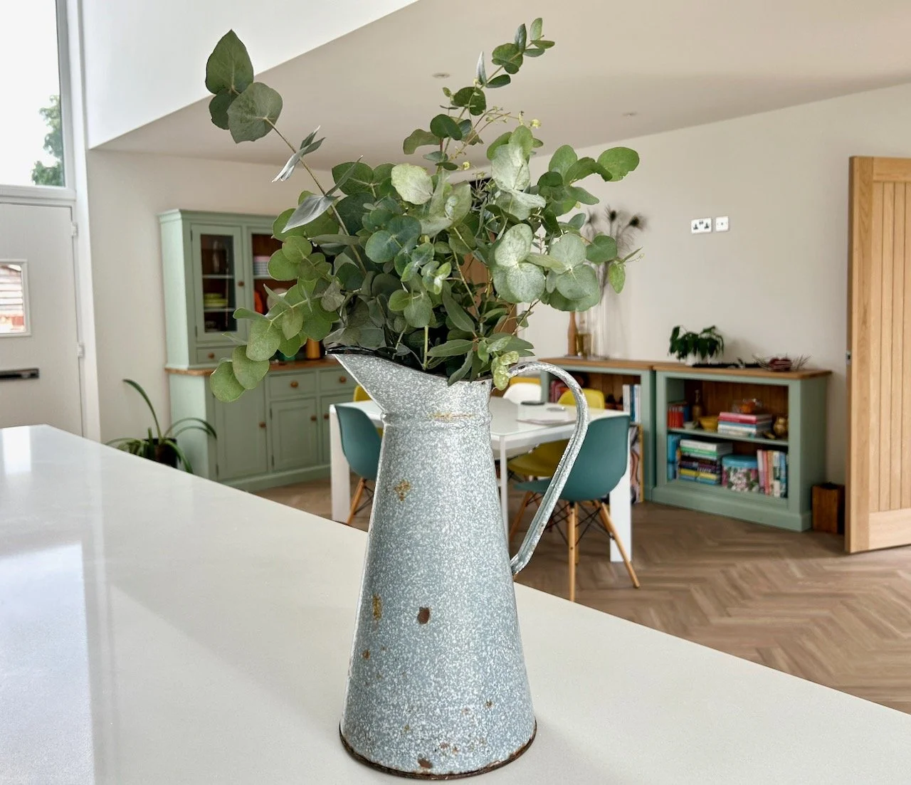 The grey mottled enamel jug filled with eucalyptus stems, on our kitchen worktop with the dining area in the background
