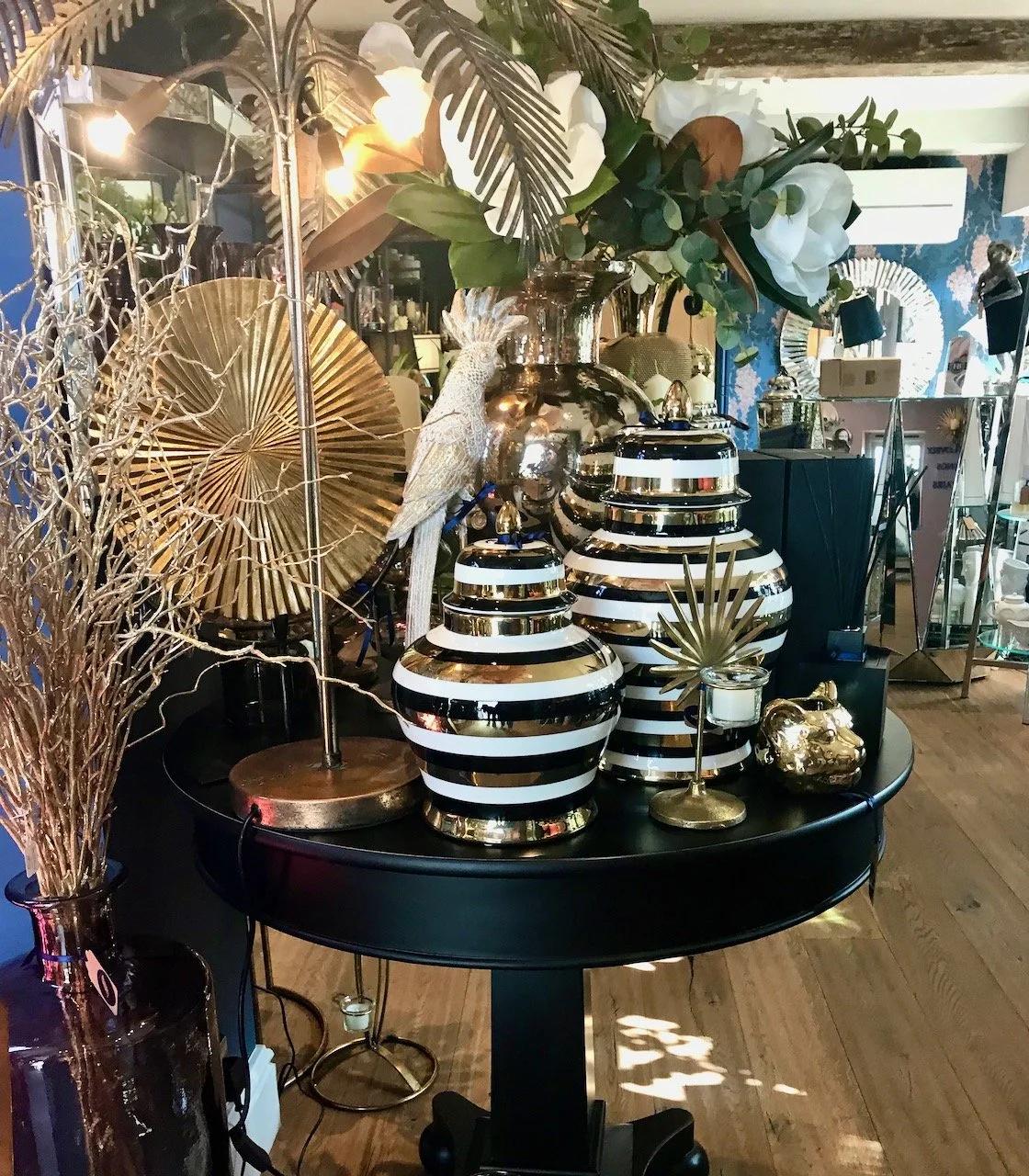 A luscious table with black gold and white striped ornate pots, gold fans, a silver cockatiel (not real) and tropical like greenery
