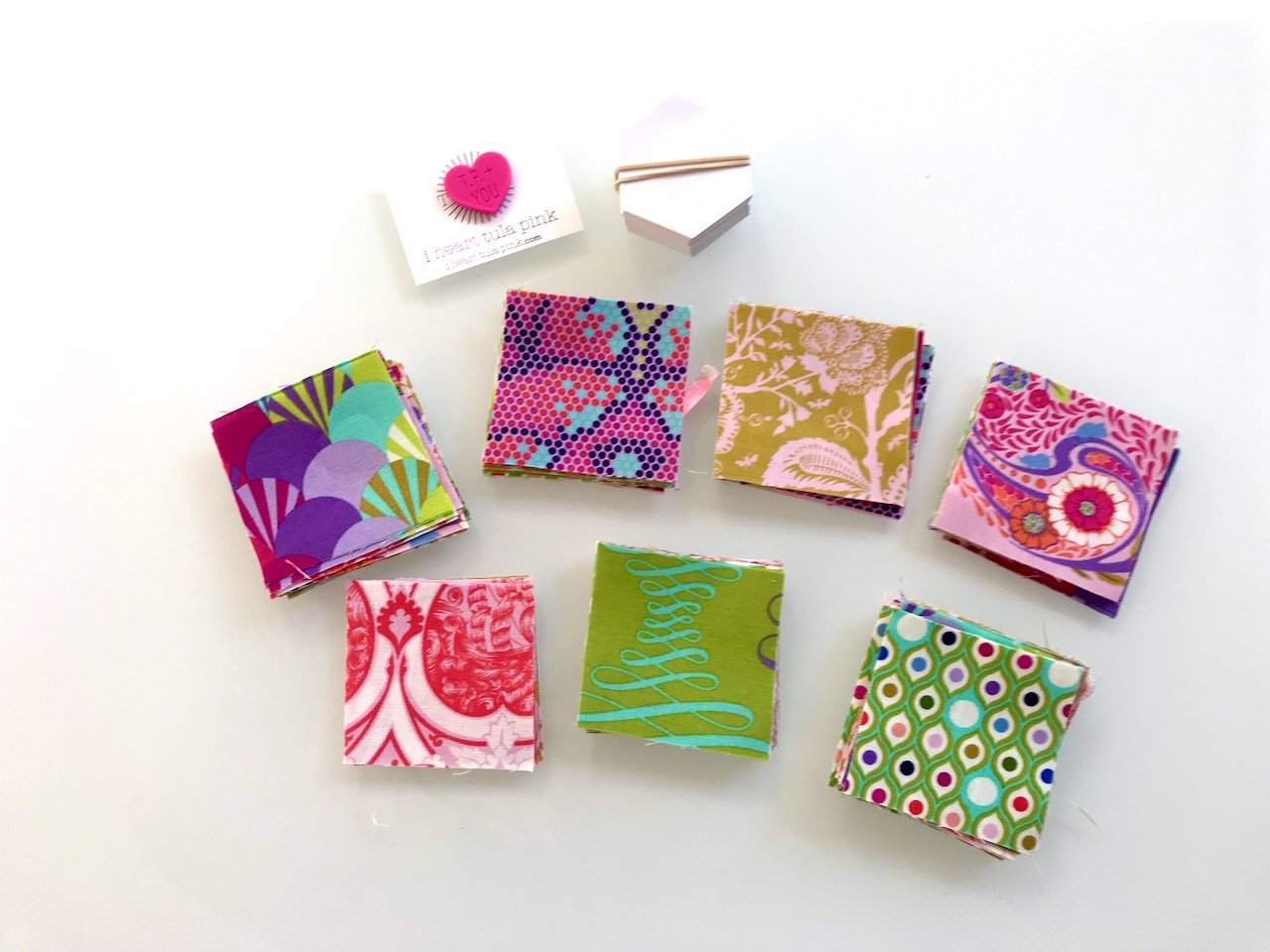 A selection of the brightly coloured fabrics in the Parisville Deja Vu Tula Pink range, a pink heart shaped magnet and white hexagonal cards for EPP