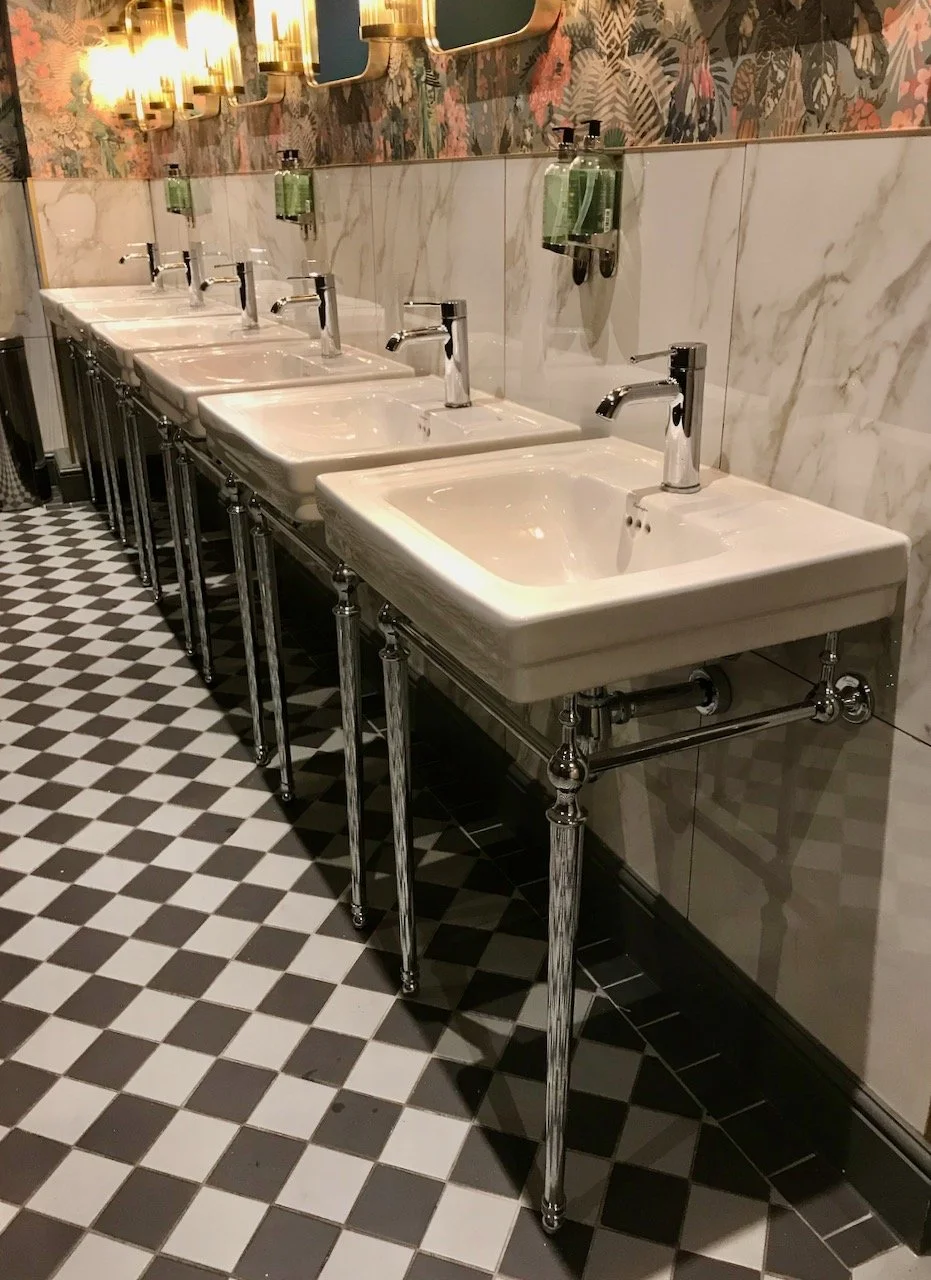A row of six white sinks on chrome legs, a black and white geometric floor