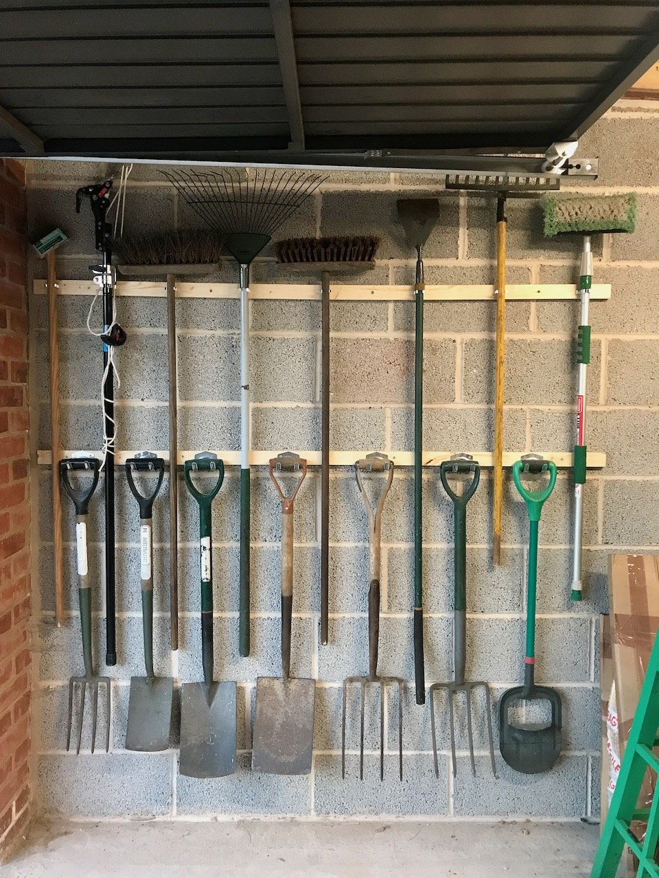 Brooms, rakes and hoes now hanging on the top wooden batten on the garage wall with spades, shovels, forks and edge cutters hanging on the lower batten