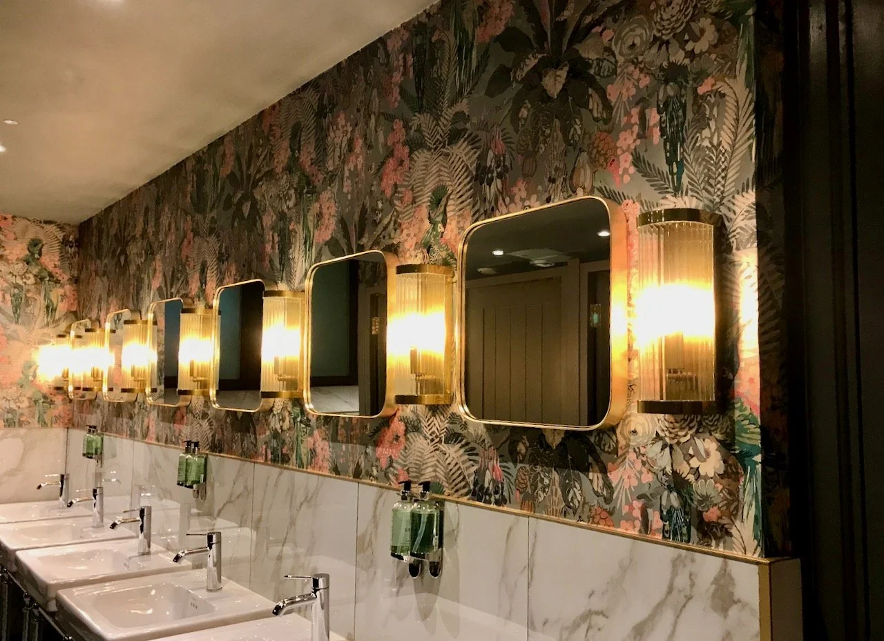 Above the sinks are rounded square mirrors with glass tube wall lights in between, set on a tropical lush wallpaer