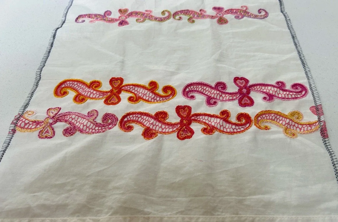 Hand embroidery in two sections - the top has a row of pastel emblems, after a break there's two rows of bolder pink and orange emblems
