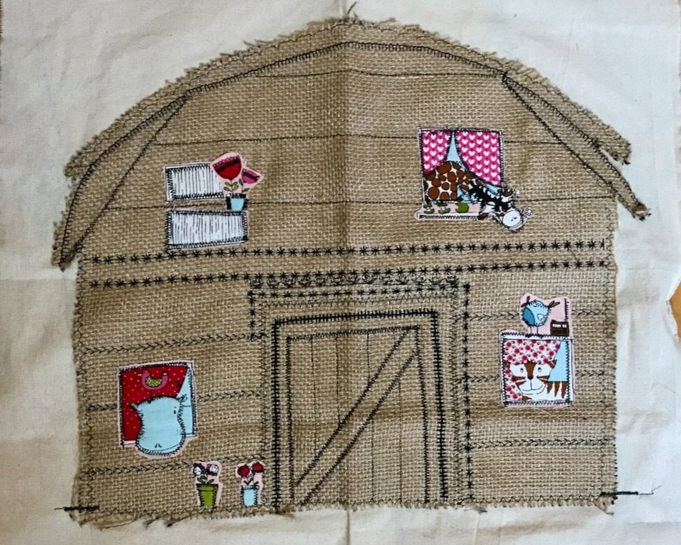 A rustic hessian barn shape on cream backing fabric.  The barn is decorated with appliqued 'windows' and uses decorative stitches to create the barn door and other panelling/decorative touches on the barn