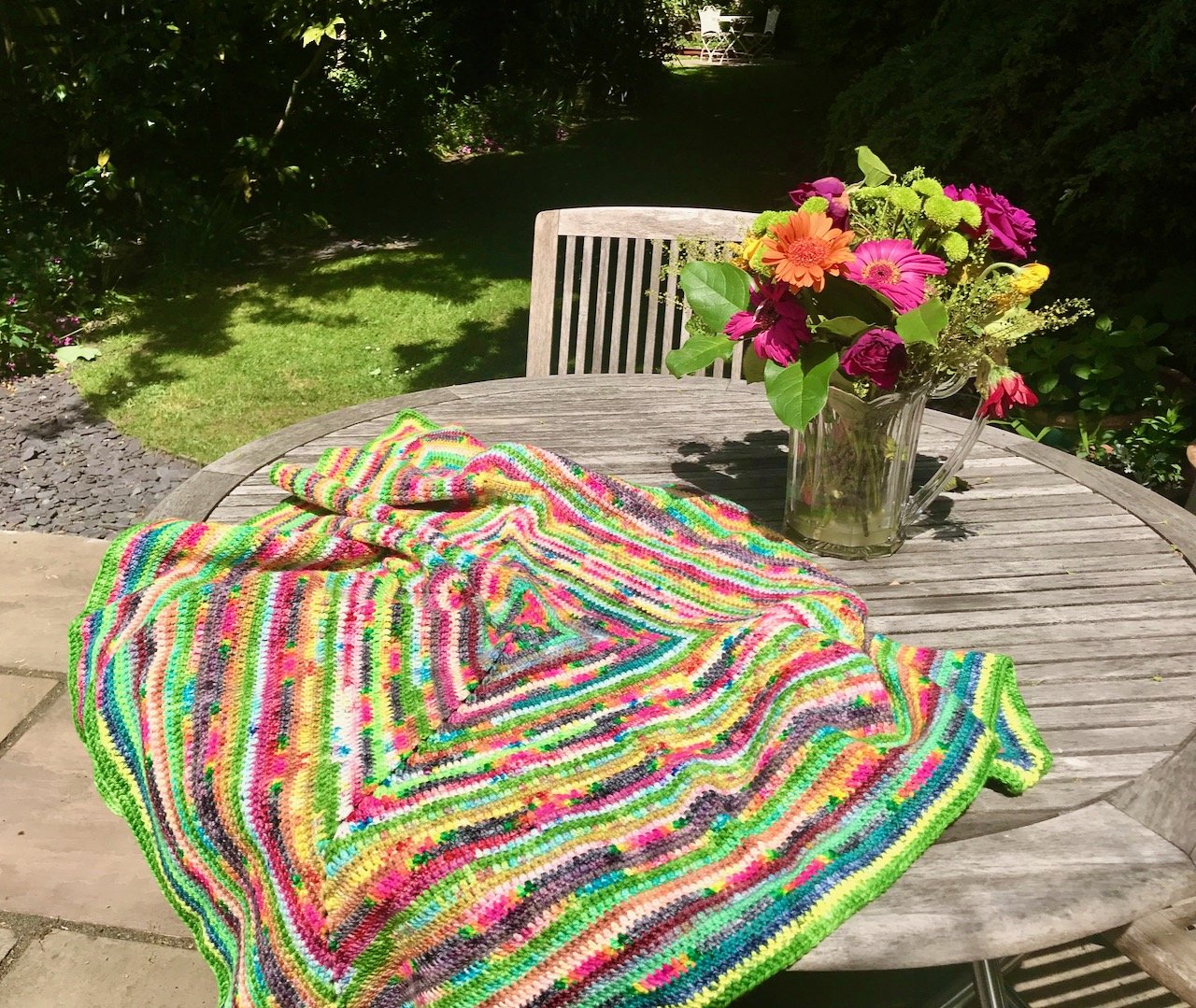 a brightly coloured crochet blanket thrown across a  garden table with a jug of brightly coloured flowers alongside
