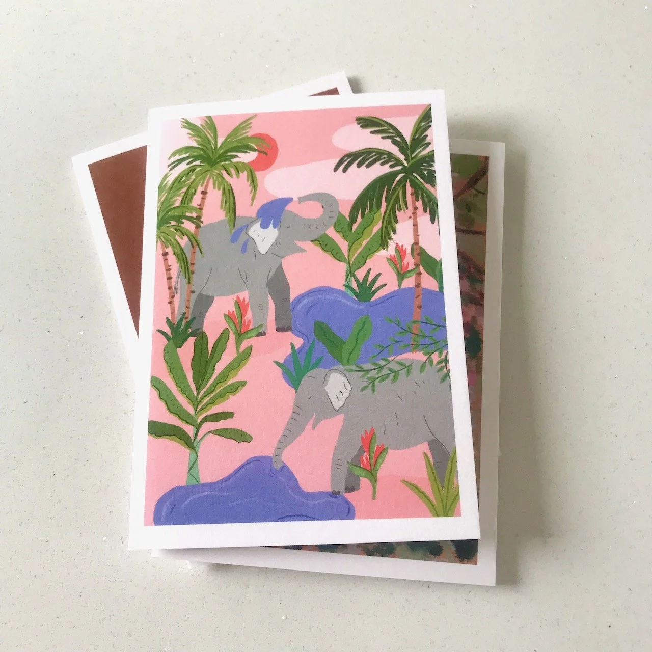 Cheerful elephants at a watering hole surrounded by palm trees, one elephant is hosing water over its head, the other has its trunk in a pool of water - all on  a cheery pastel pink background