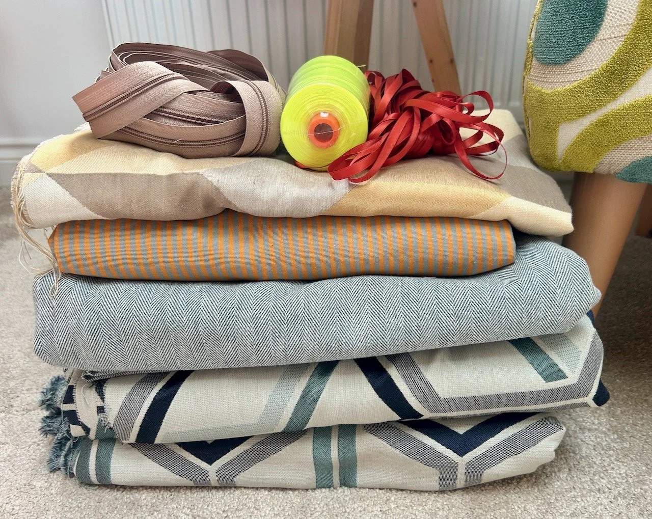A pile of five folded fabric lengths, topped with a roll of continuous zipper, neon thread and some rust coloured ribbon
