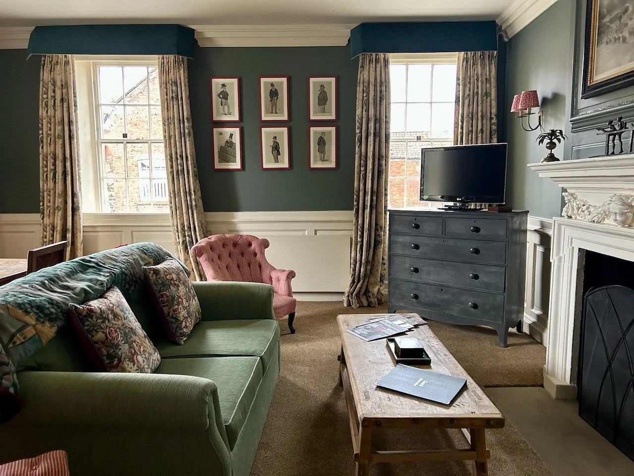 A traditional styled room with a sofa in front of the fire, two windows traditionally dressed and a series of six portraits hanging between them