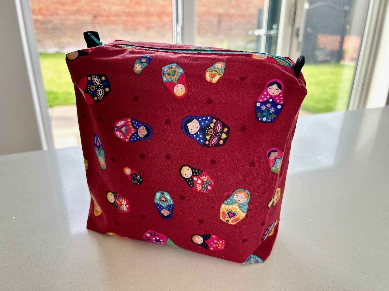 A tall rectangular pouch in burgundy fabric decorated with russian doll motifs