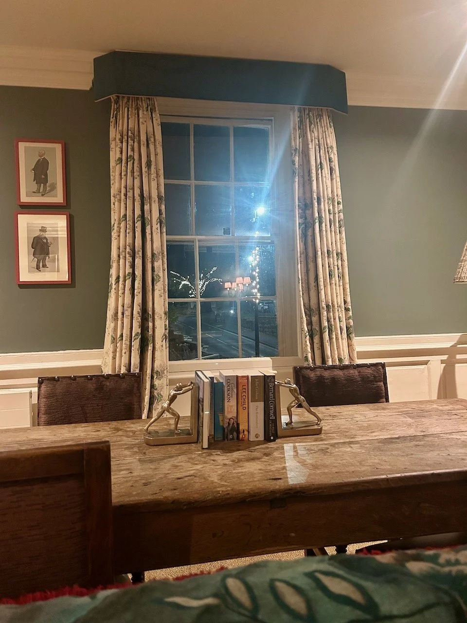 A large wooden and weathered table in the foreground with reading books and bookends, another window with the same traditional dressing and two further portraits framed on the wall