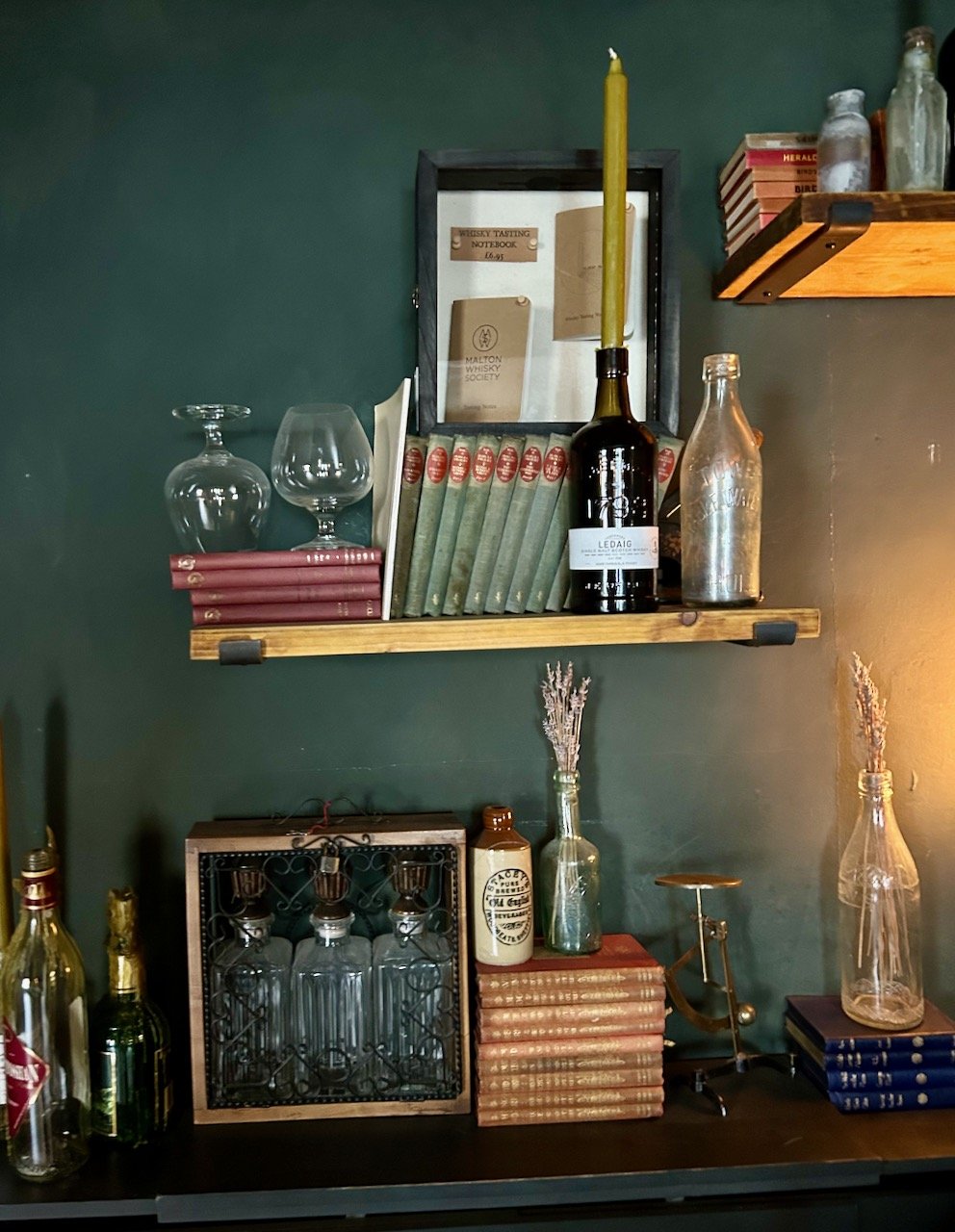 The focus is a wooden floating shelf with books, brandy glasses and two bottles.  Below it on the wooden unit top are more books and more decanter type vessels