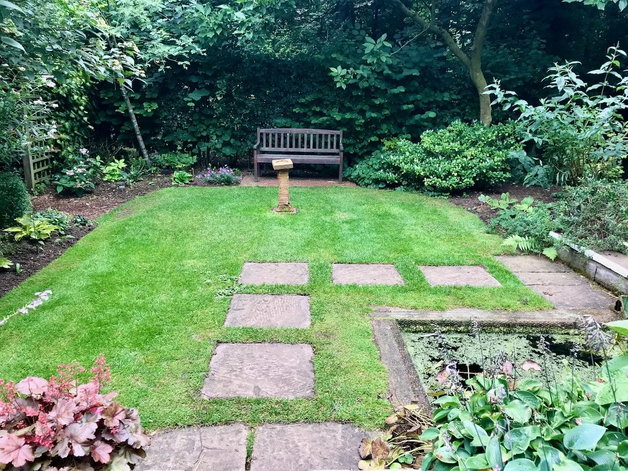 a bench in the background with hedges/greenery behind.  A central lawn with square pavers as stepping stones forming a right angled path around the hidden pond