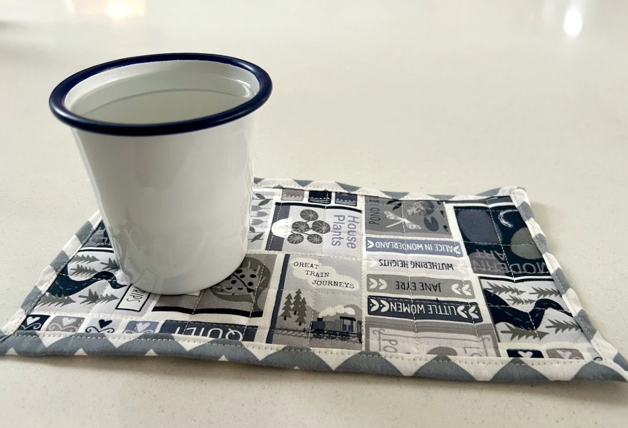 A two cup by the sink mat made from grey patterned fabric with an enamel tumbler currently on the mat