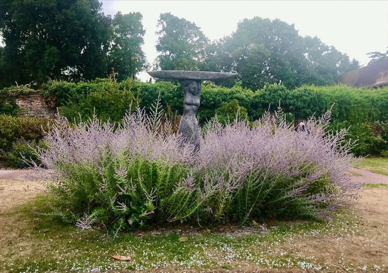 A stone statue surrounded by purple flowering plants set in a grass border