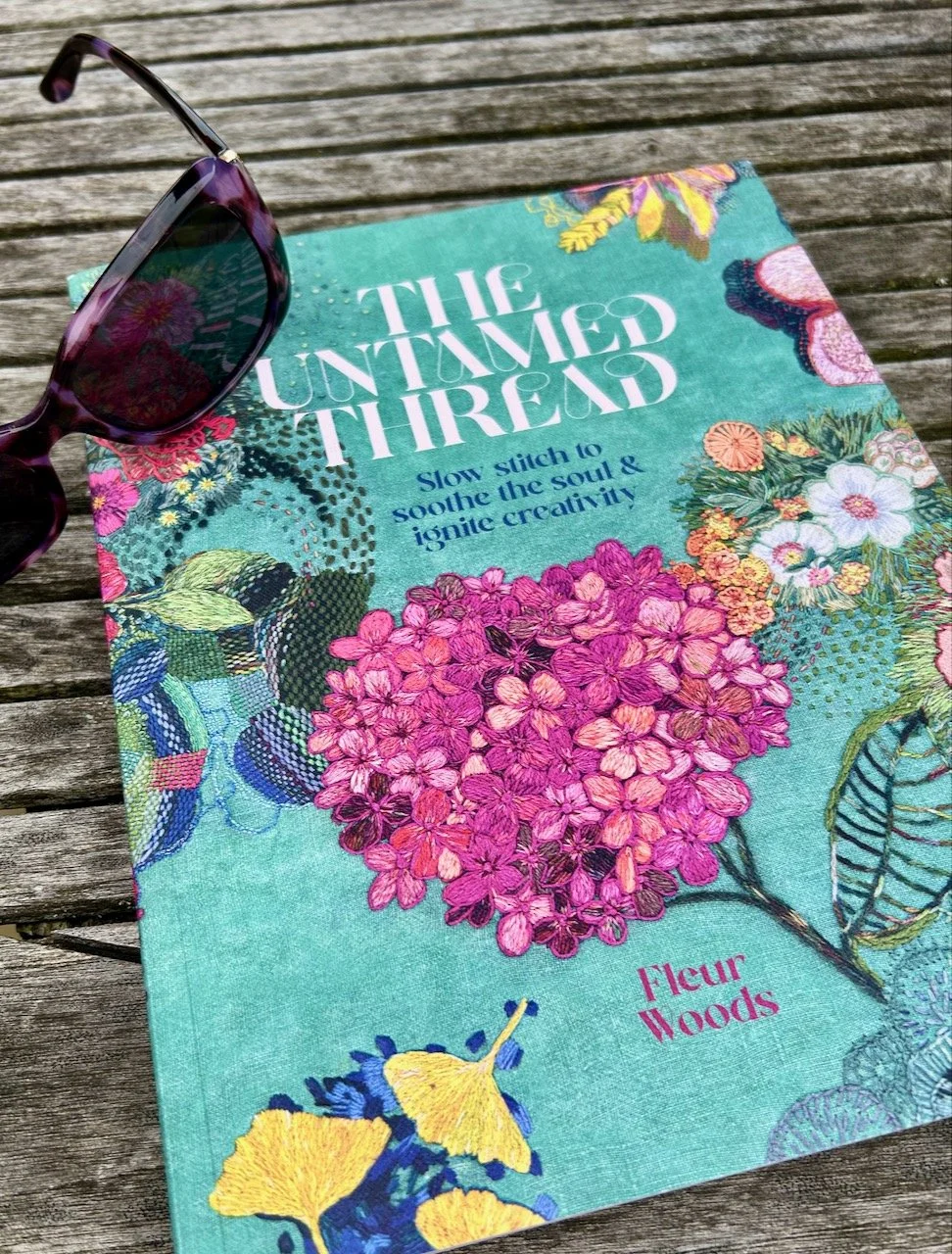The Untamed Thread book by Fleur Woods on a wooden garden table with sunglasses