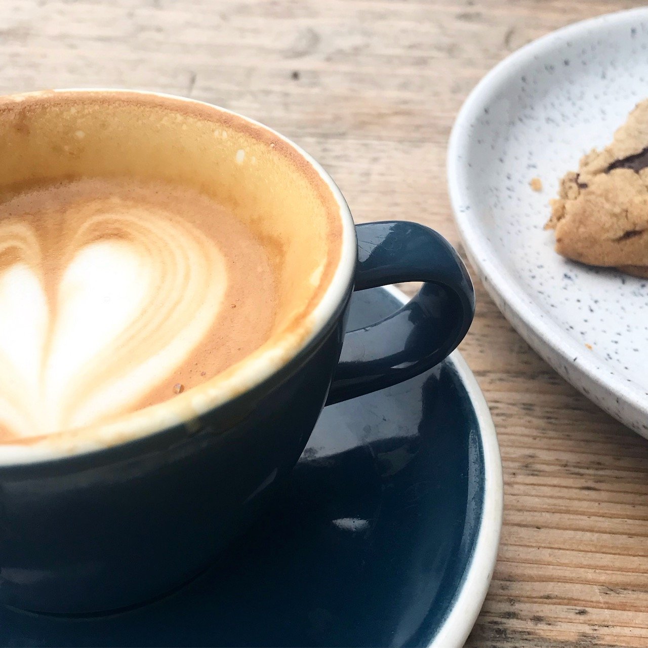 A flat white - with heart shaped coffee art - in a navy cup with part of a chocolate chip cookie on a speckled plat alongside