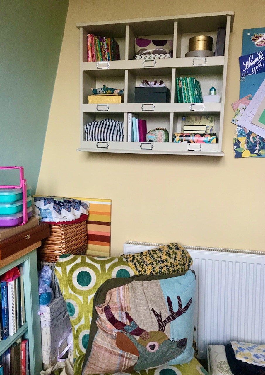 A longer shot of the shelves above showing more of the room with an arm(less) chair in front and bookshelves to the left.  A real cosy place to sit and relax (and craft!)