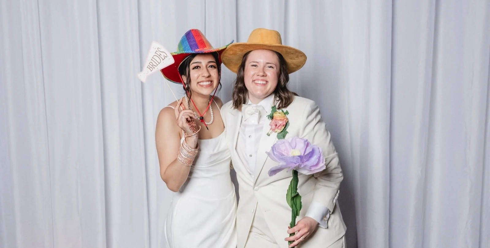 Two women smiling, wearing playful hats, one with a rainbow sombrero labeled 'BRIDE' and the other with a straw hat, posing together at a wedding celebration, one holding a large purple paper flower.