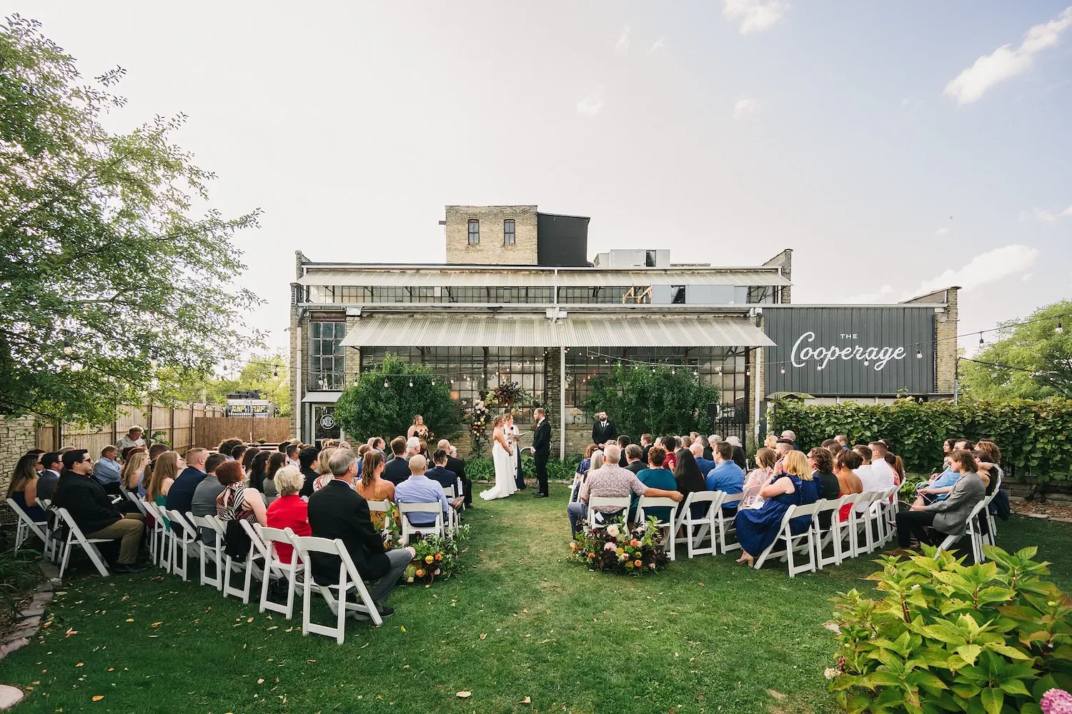 An outdoor wedding ceremony with a bride and groom exchanging vows in front of guests seated on white chairs on a grassy lawn, in the back courtyard of the Cooperage, an event space near Lake Michigan in Milwaukee (connected to Boone and Crocket)!