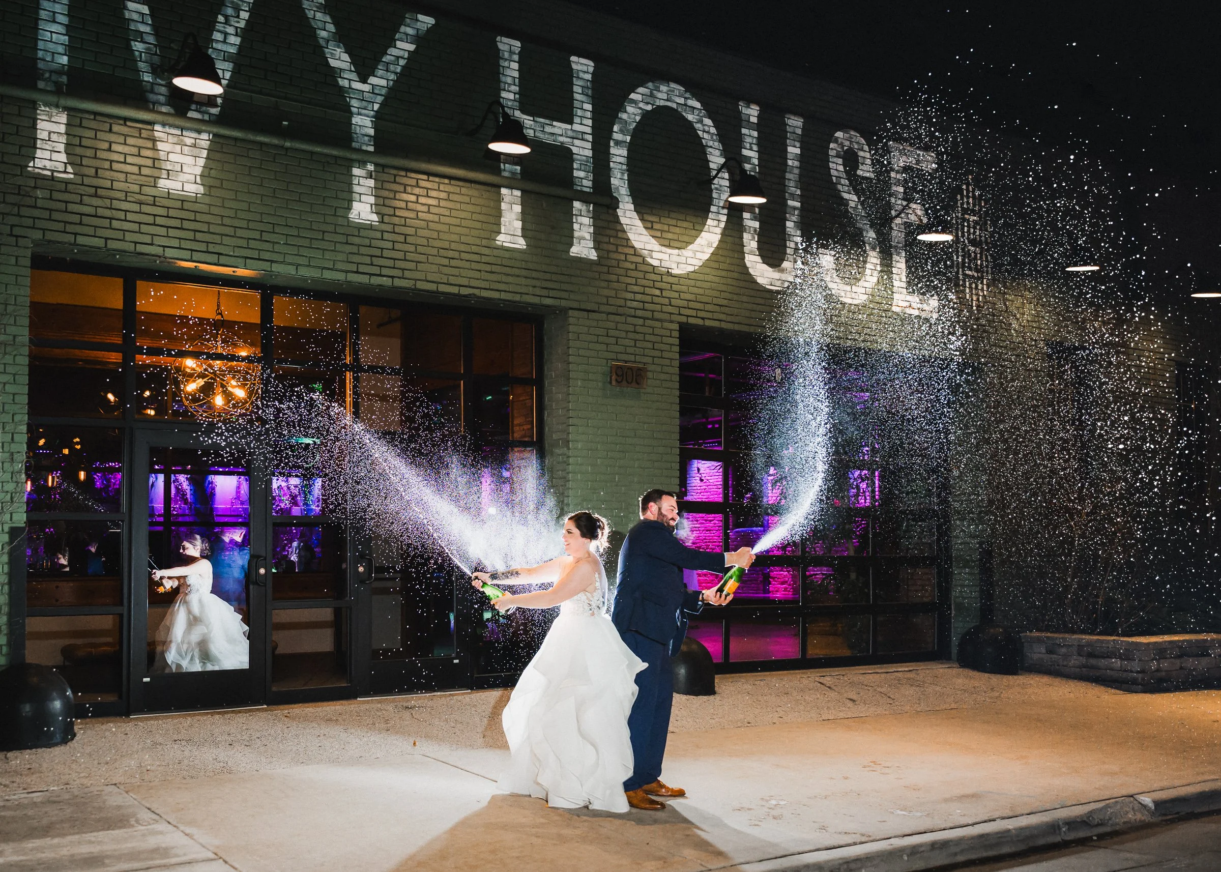 A bride and groom celebrating outside a venue named 'The Yard House' with champagne bottles spraying fireworks in front of a brick building during nighttime.