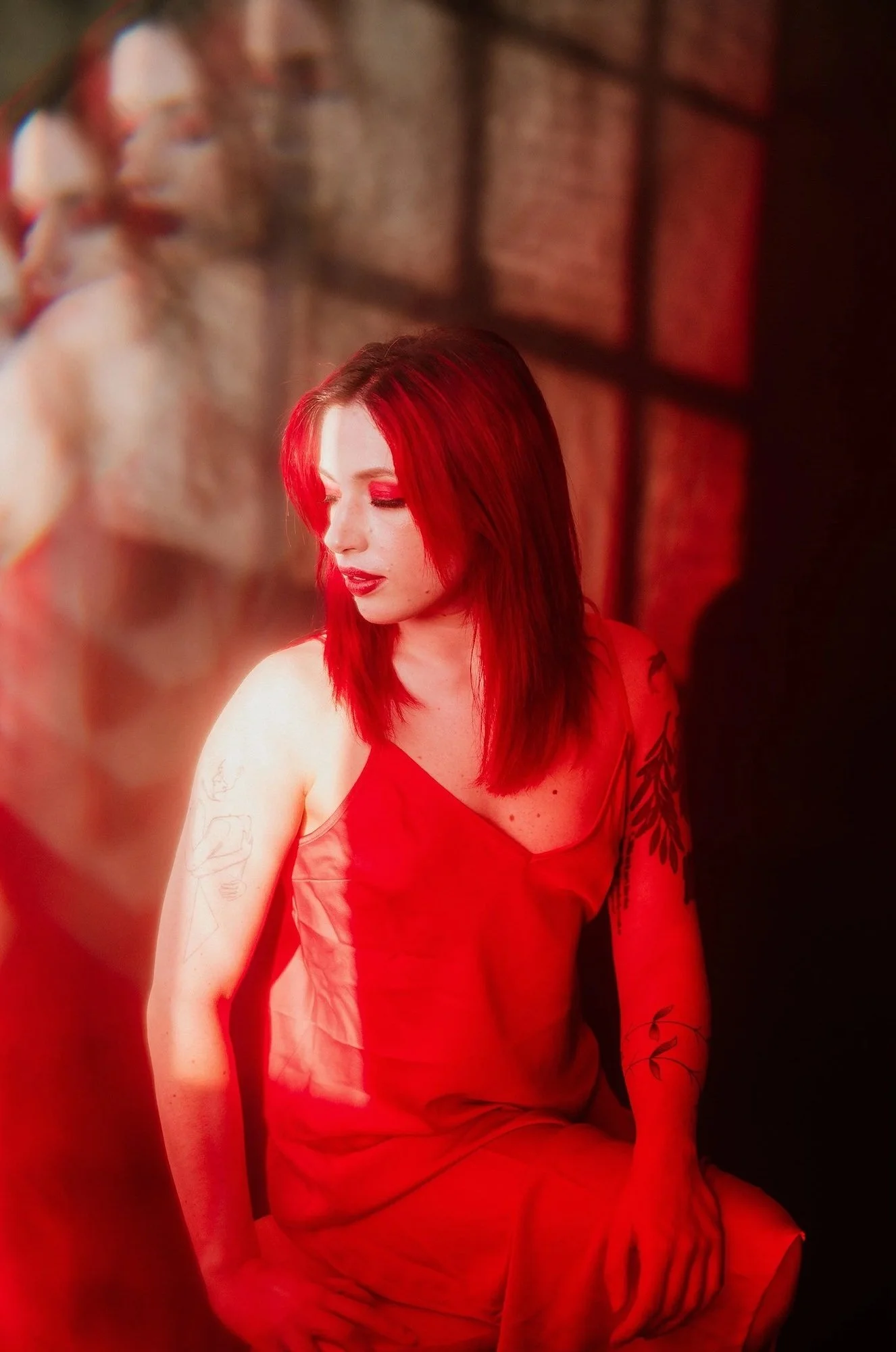 A woman with red hair and tattoos wearing a red dress sitting in a dimly lit room with red lighting.