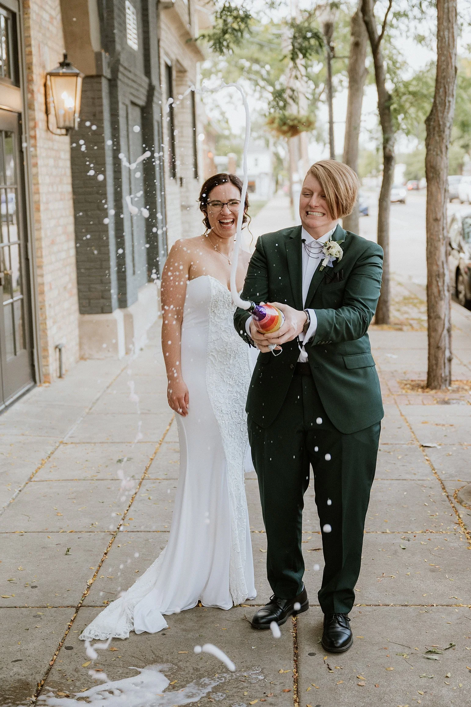 A couple celebrating with a champagne spray on a sidewalk, with one in a white wedding dress and the other in a green suit.