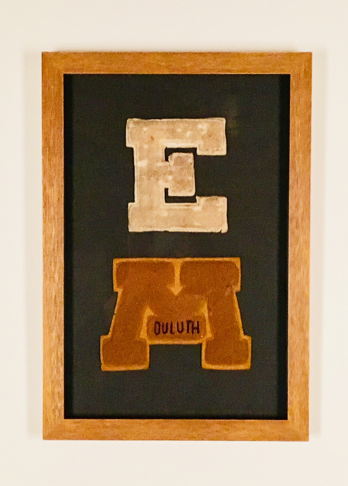 Varsity letters sewn to charcoal linen and shadowboxed in ochre frame.
