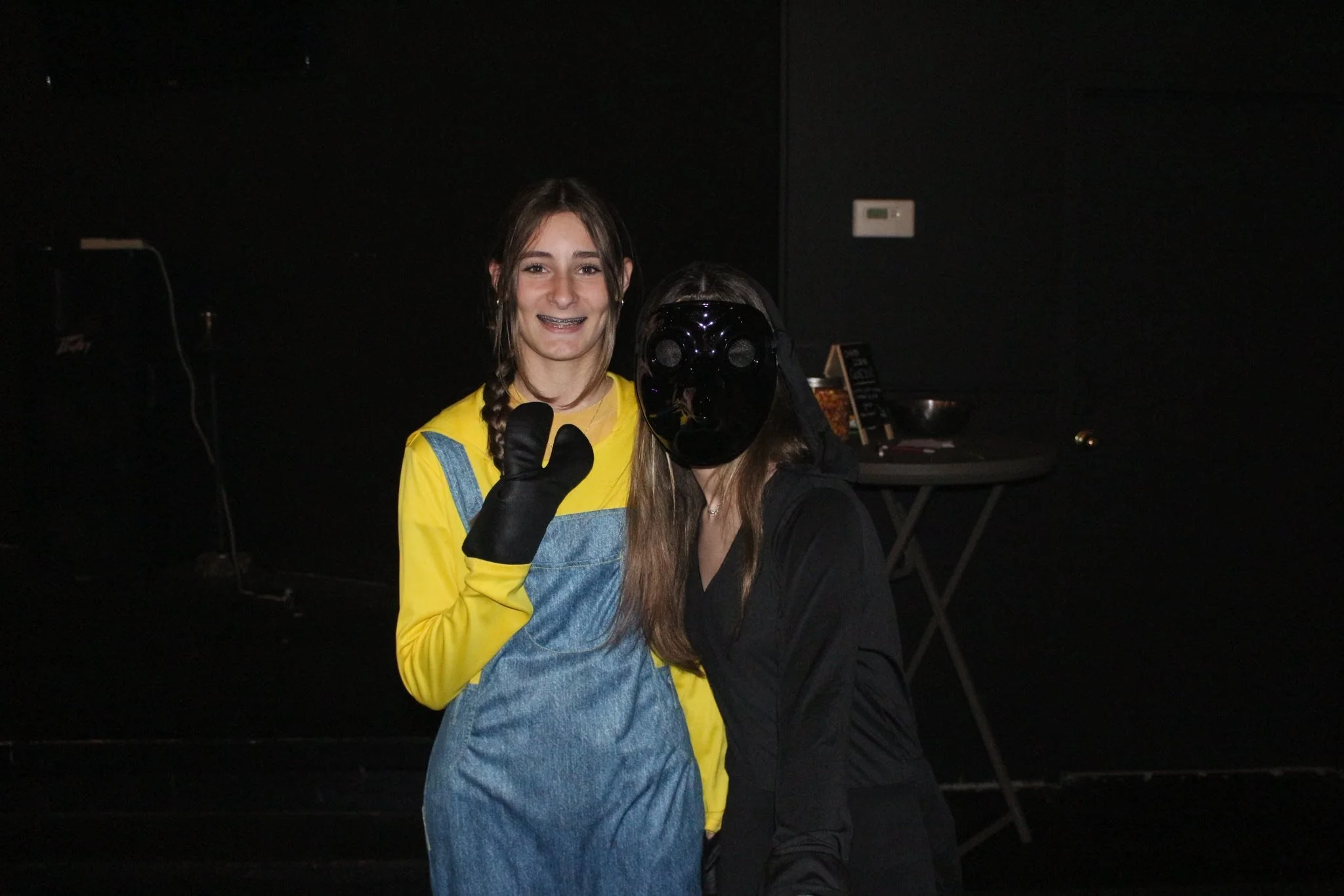 Two women standing together in a dark room, one smiling and waving while wearing a yellow shirt, denim dress, and black glove, the other wearing a black outfit with a black mask covering their face.