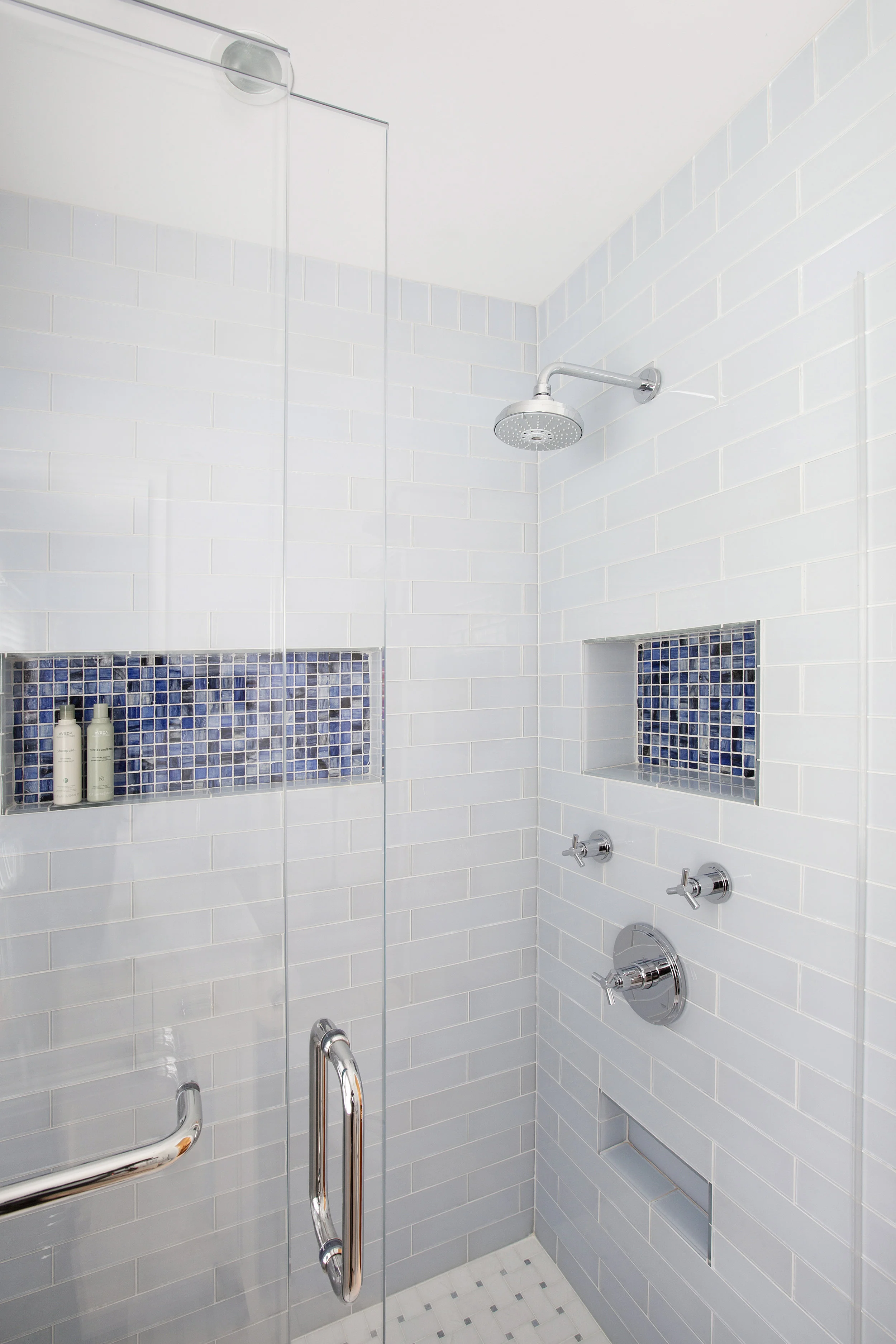 Blue glass tile provides a splash of color in shower niches; the flooring is white marble with blue azul accents. A small niche below the faucets provide a footrest for shaving legs. 