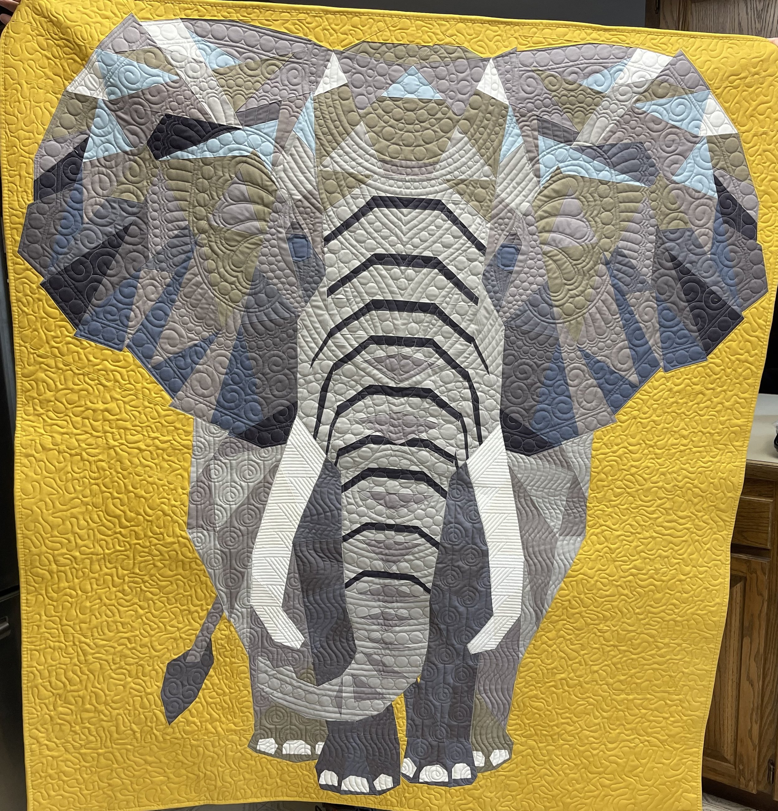 A quilt of an elephant facing forward on a gold background. The elephant in made of grey and blue patterned fabric.