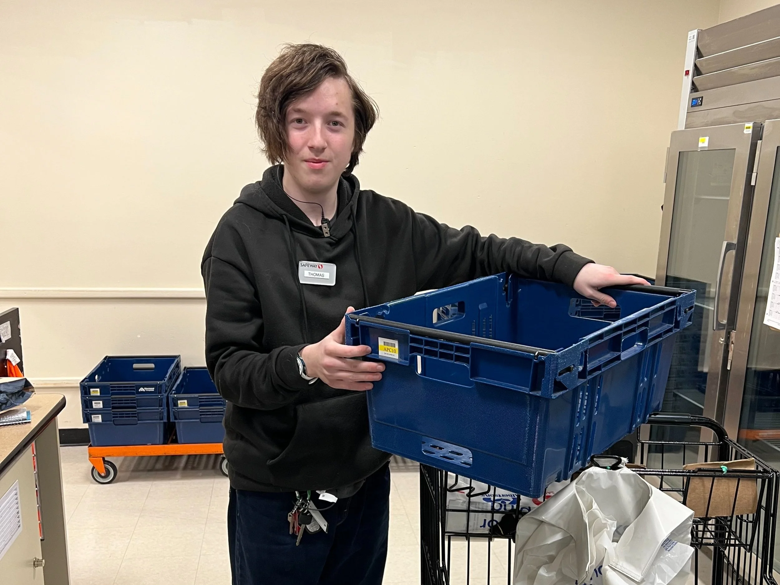 Thomas Nelson poses for a picture at work. He wears a black hoodie with his Safeway nametag. He is holding a plastic bin used for internet orders.