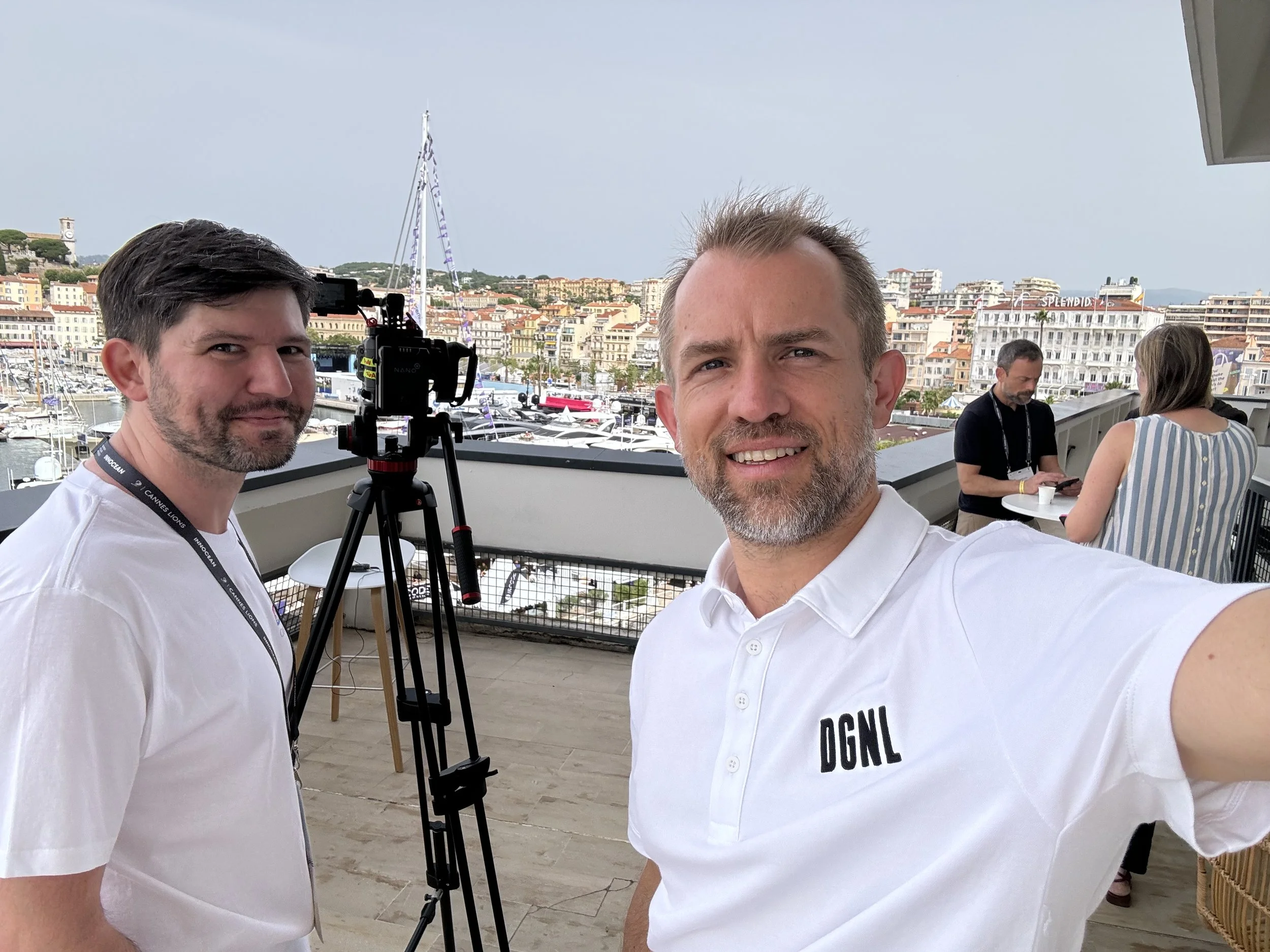 Two men taking a selfie on a balcony overlooking a marina with yachts and a cityscape in the background. One man is holding the camera and has a short beard, wearing a white polo shirt with 'DGLN' embroidered. The other man, standing beside him, has 