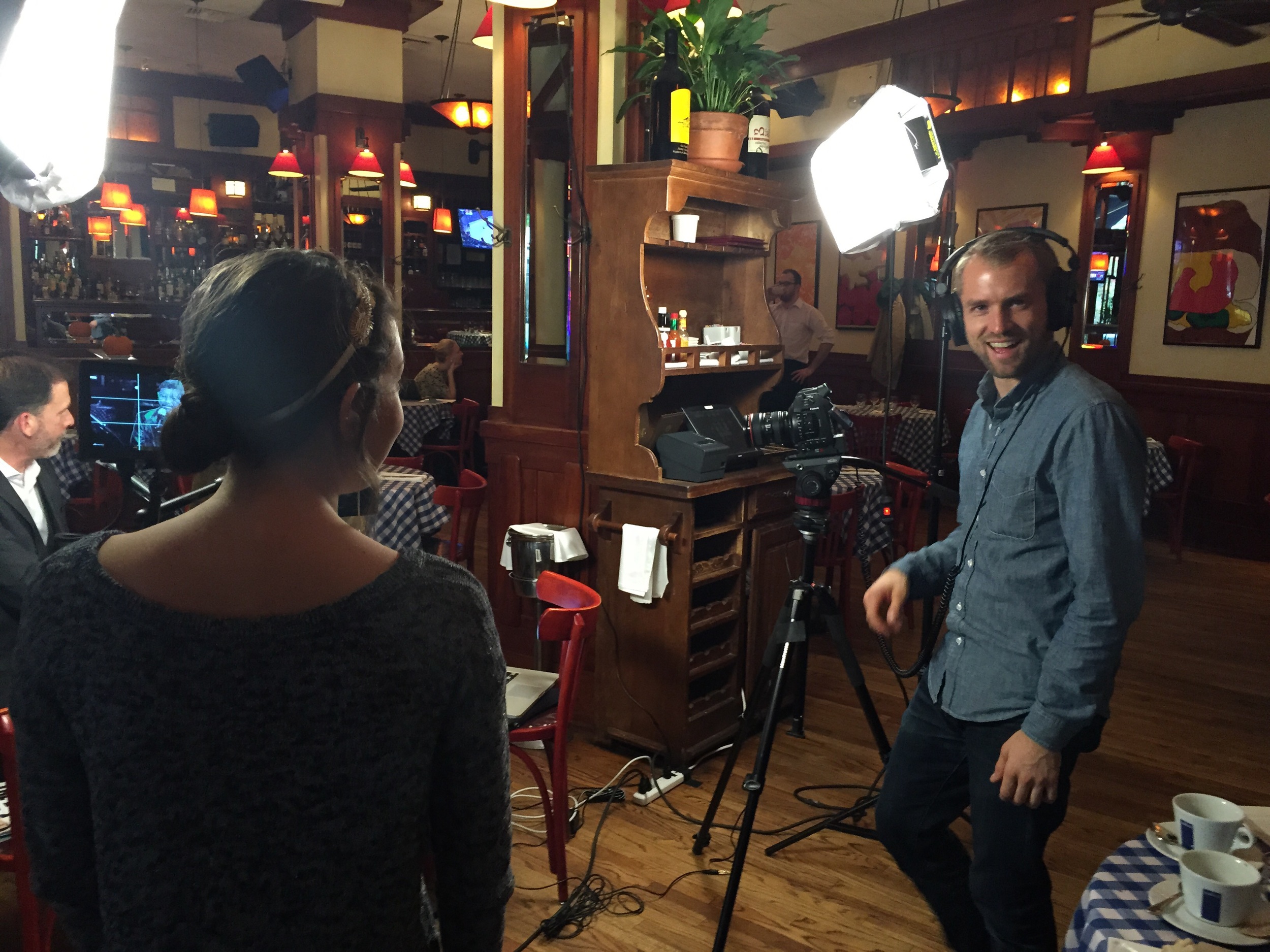 A woman being filmed at a restaurant with a cameraman and professional lighting. The woman is seen from behind, and the cameraman is smiling at the camera.