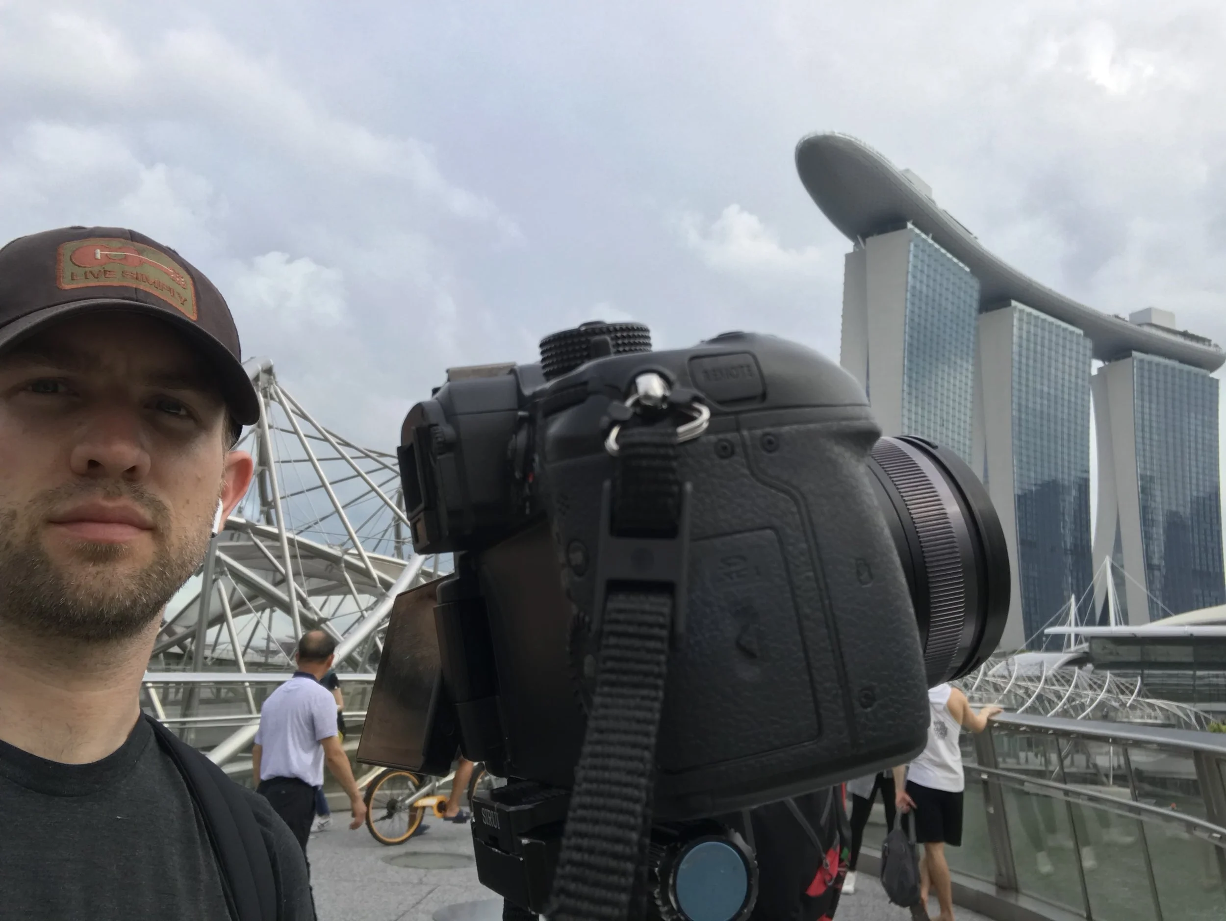 A man with a camera and earbuds standing outdoors near Marina Bay Sands hotel and the ArtScience Museum in Singapore, with cloudy skies.