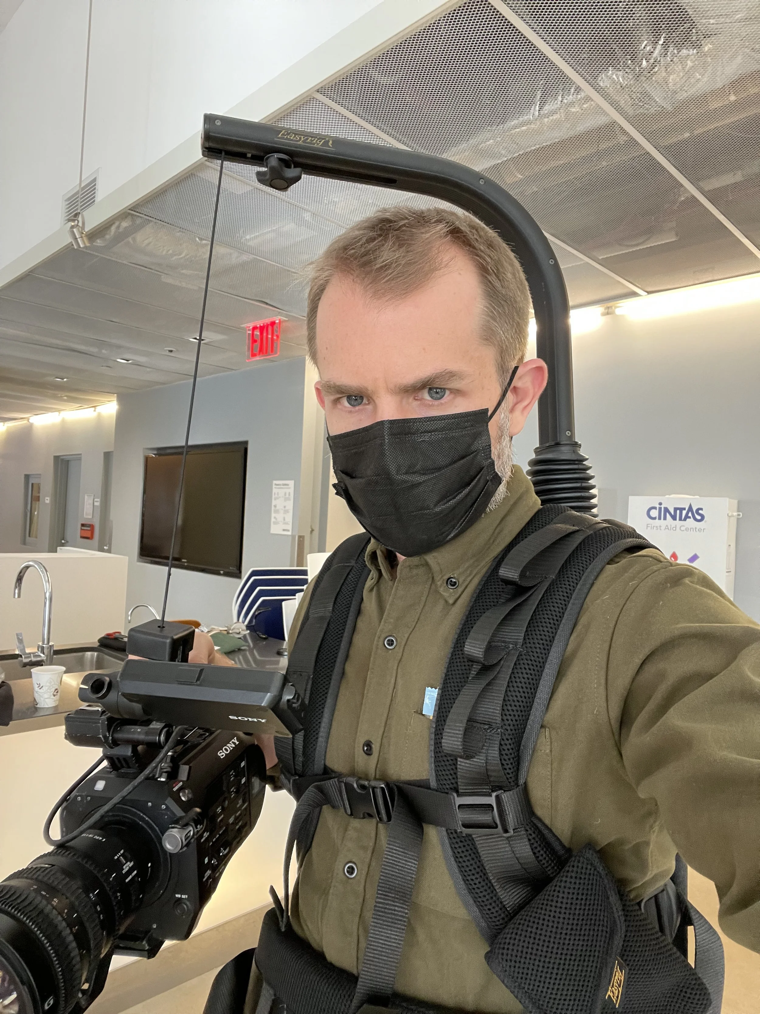 A man wearing a black face mask, olive green button-up shirt, and carrying a camera rig with a Sony camera, standing in an airport or office environment.