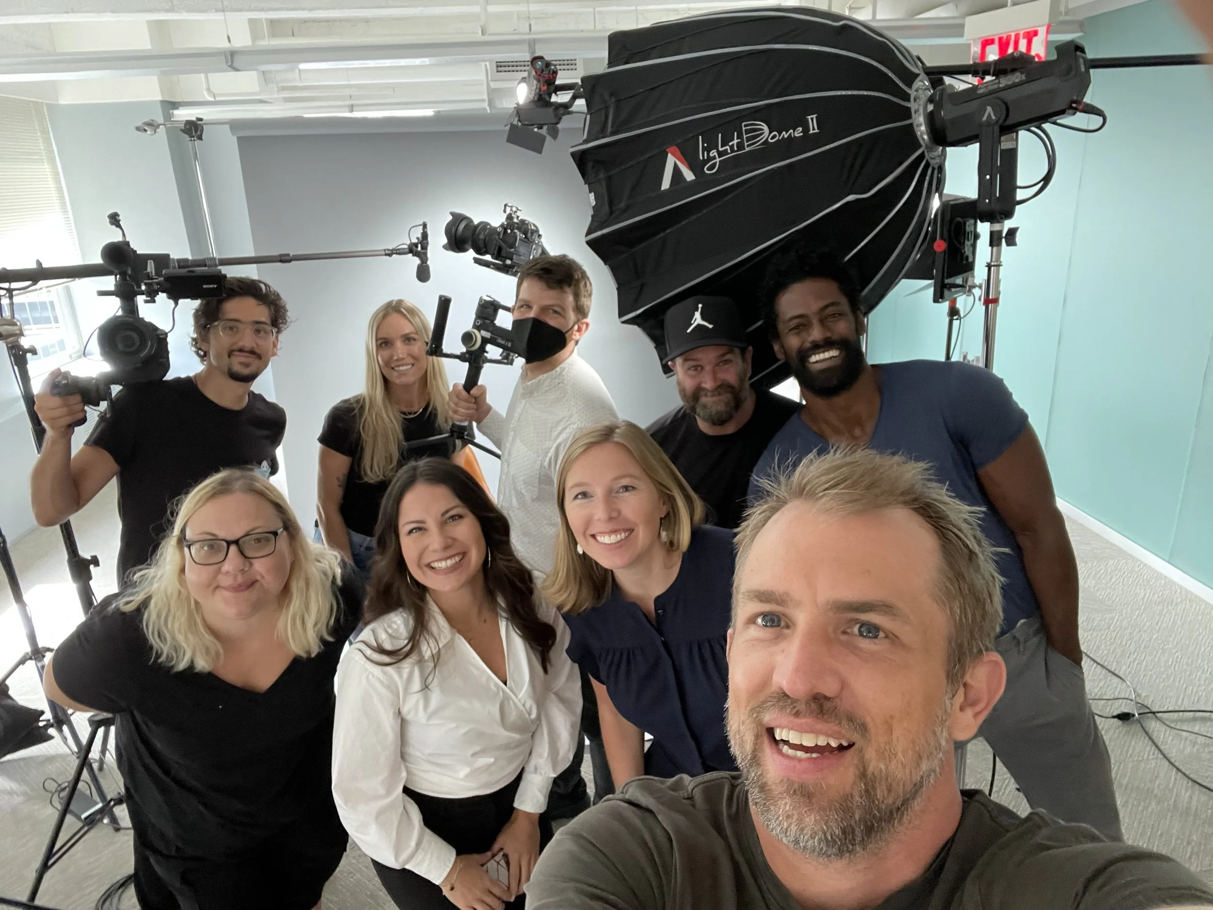 Group of nine diverse people smiling and taking a selfie in a TV or film production studio, with filming equipment including cameras, microphonoes, and lighting gear around them.
