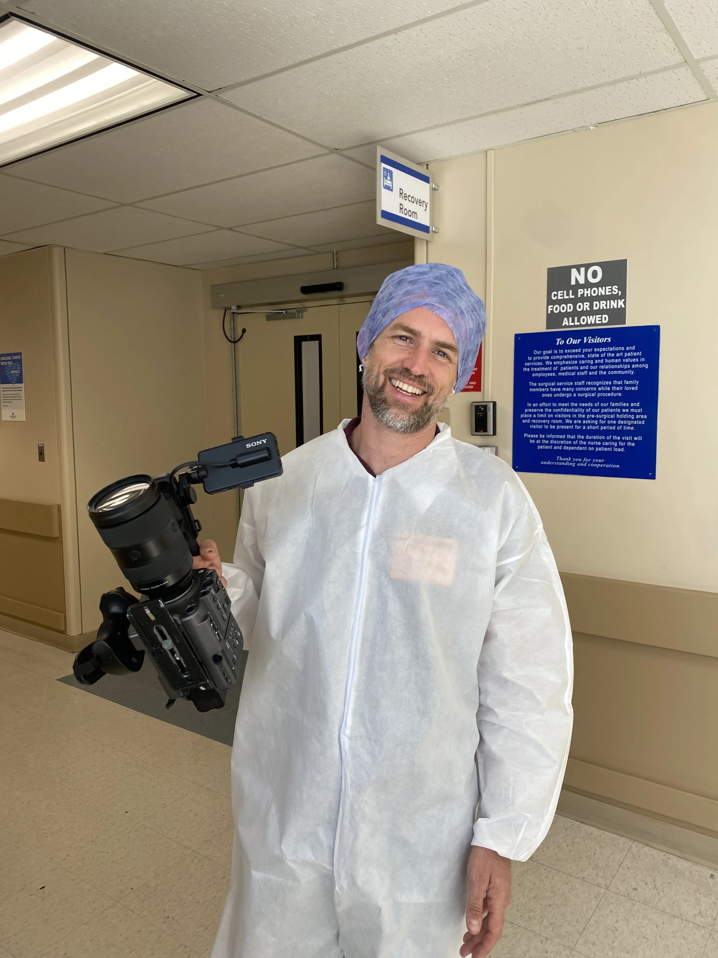 A smiling man in medical scrubs and a hairnet holding a video camera inside a hospital corridor near a sign that reads 'Recovery Room'.