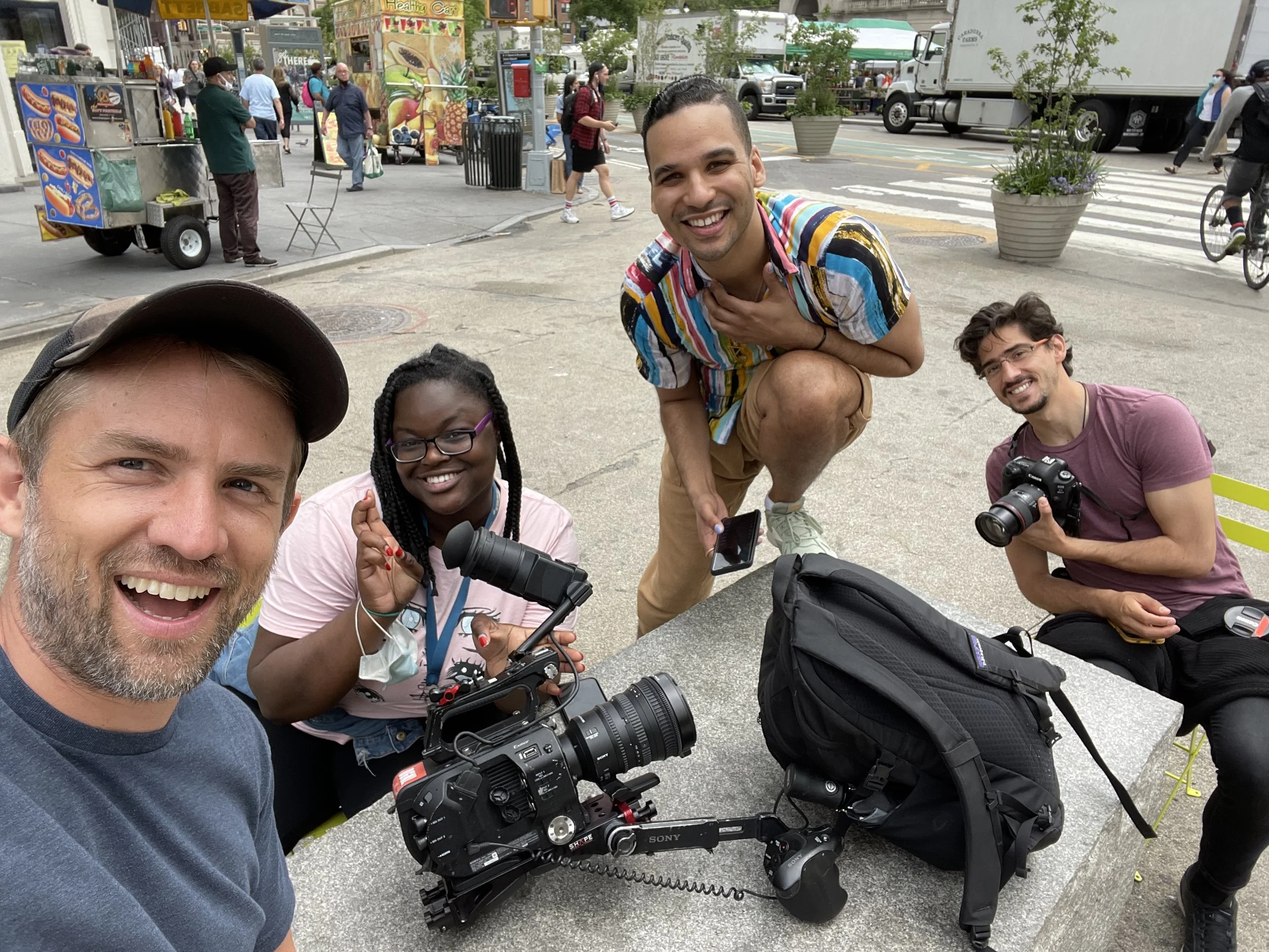 A group of five diverse people taking a selfie and smiling at an outdoor area with food carts, pedestrians, and vehicles in the background. They are sitting and standing around a concrete table with camera equipment and backpacks.