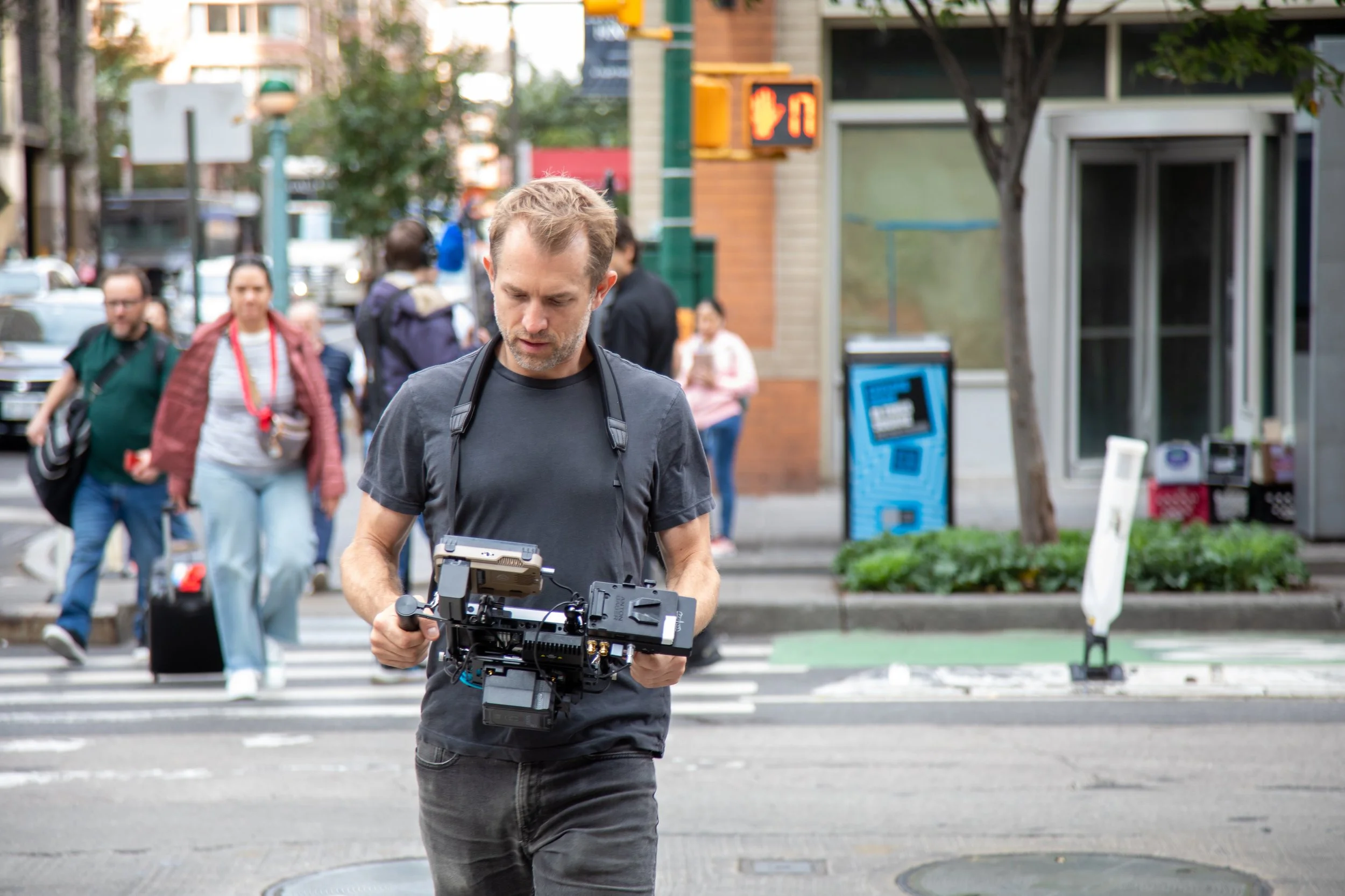 A man is walking across the street holding a professional camera stabilizer or gimbal, with people and city buildings in the background.