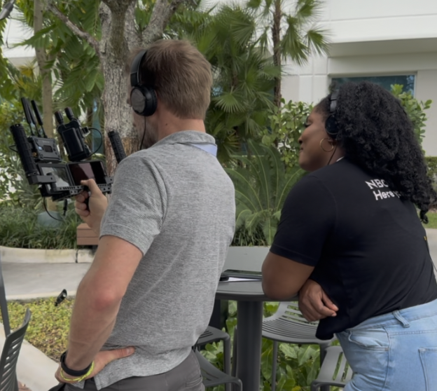 Two people working on a film set outdoors, one adjusting a camera rig and the other observing, with greenery and plants in the background.