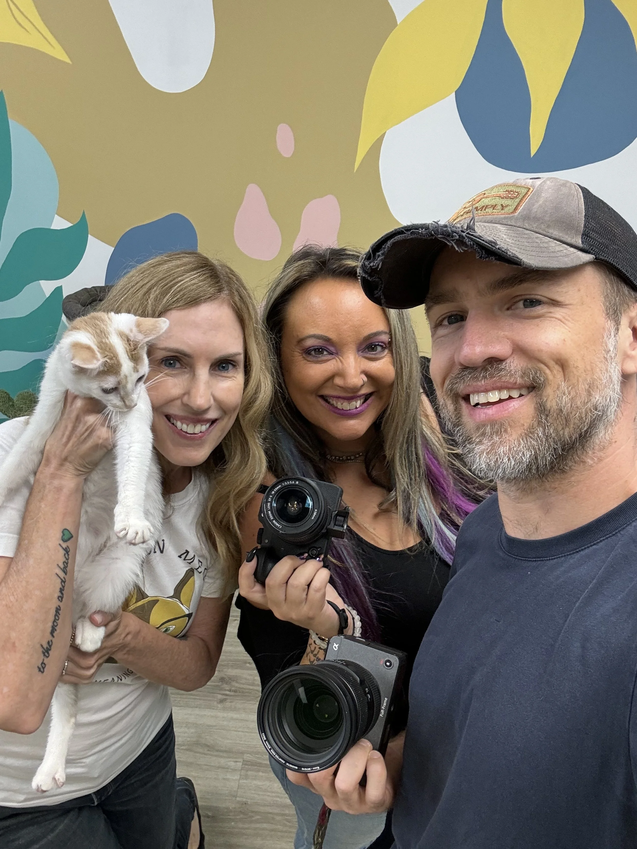 Three smiling people, two women and one man, taking a selfie indoors with a colorful, abstract wall mural in the background. The woman on the left is holding a white and orange cat, and the woman in the middle has purple hair and is holding a camera.
