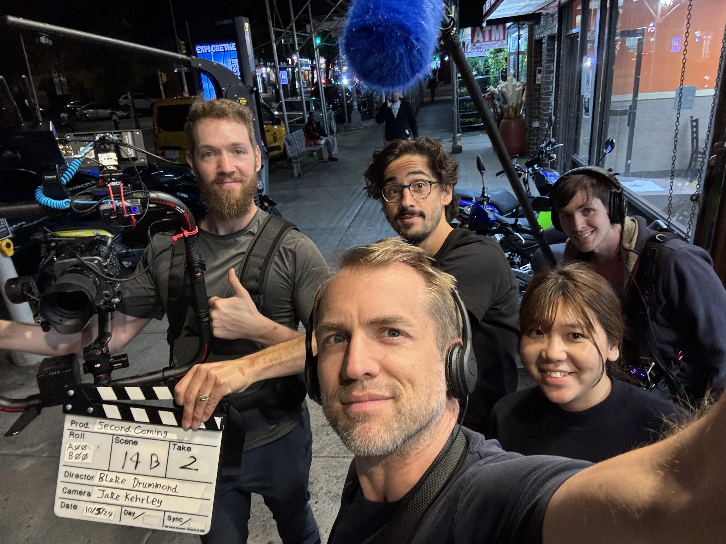 A group of five filmmakers on a city street at night, taking a selfie. One person holds a clapperboard, and another has a professional camera. They are surrounded by motorcycles and illuminated storefronts.