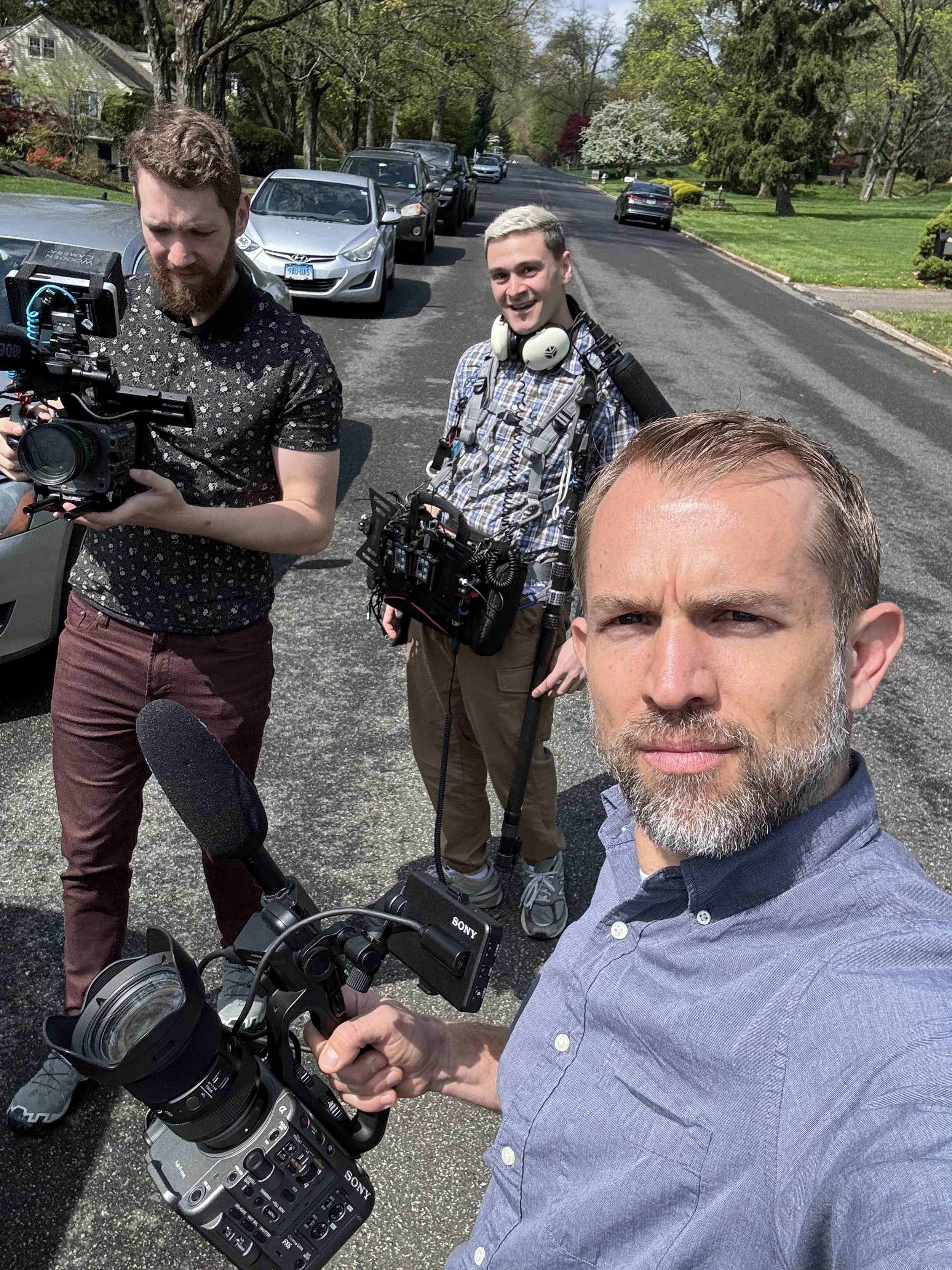 Three men standing on a residential street, two holding professional video cameras and one taking a selfie, with parked cars and trees in the background.