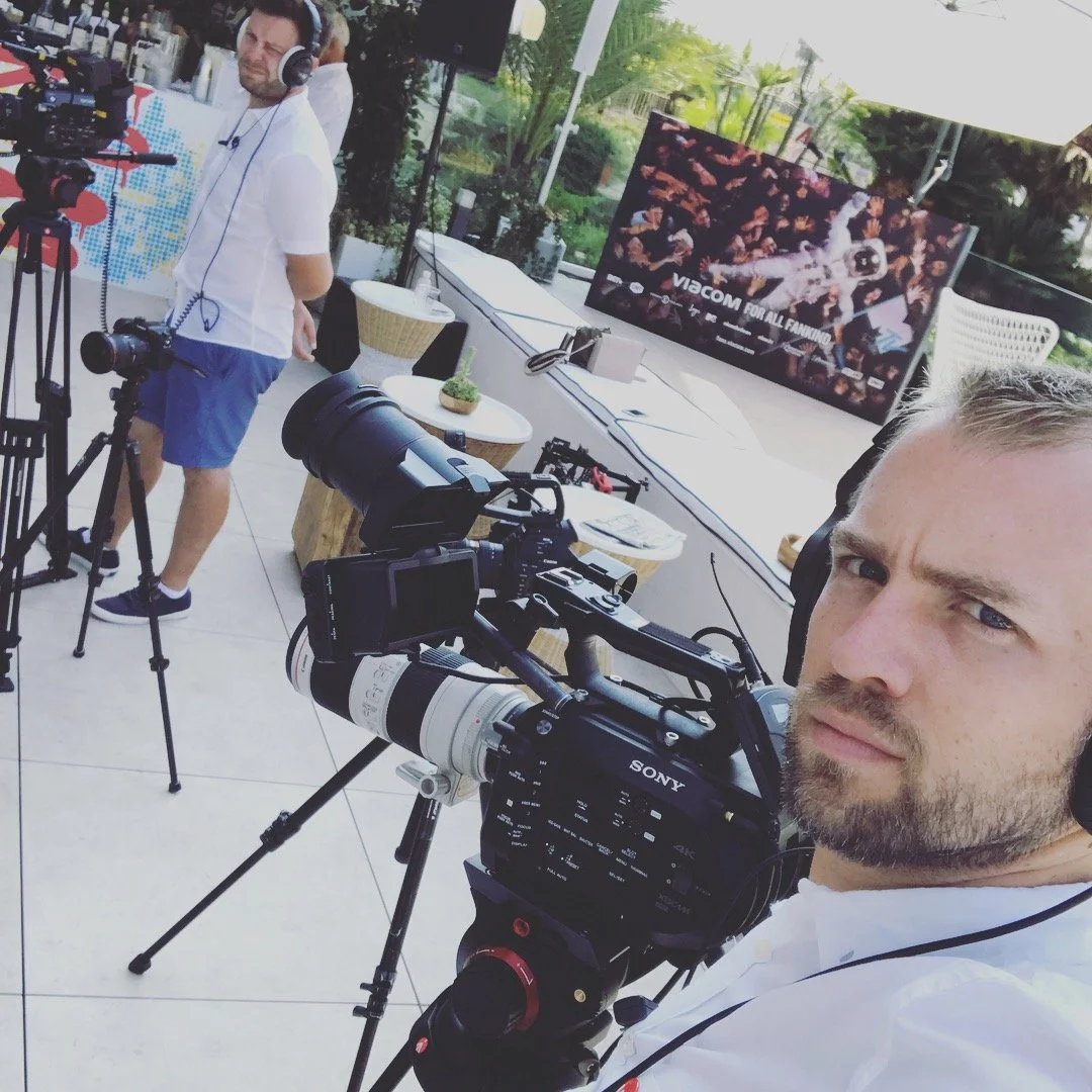 A man with a beard and white shirt taking a selfie at an outdoor event with video equipment, a person adjusting a camera, and a television displaying a Rocket League match on a patio or terrace.