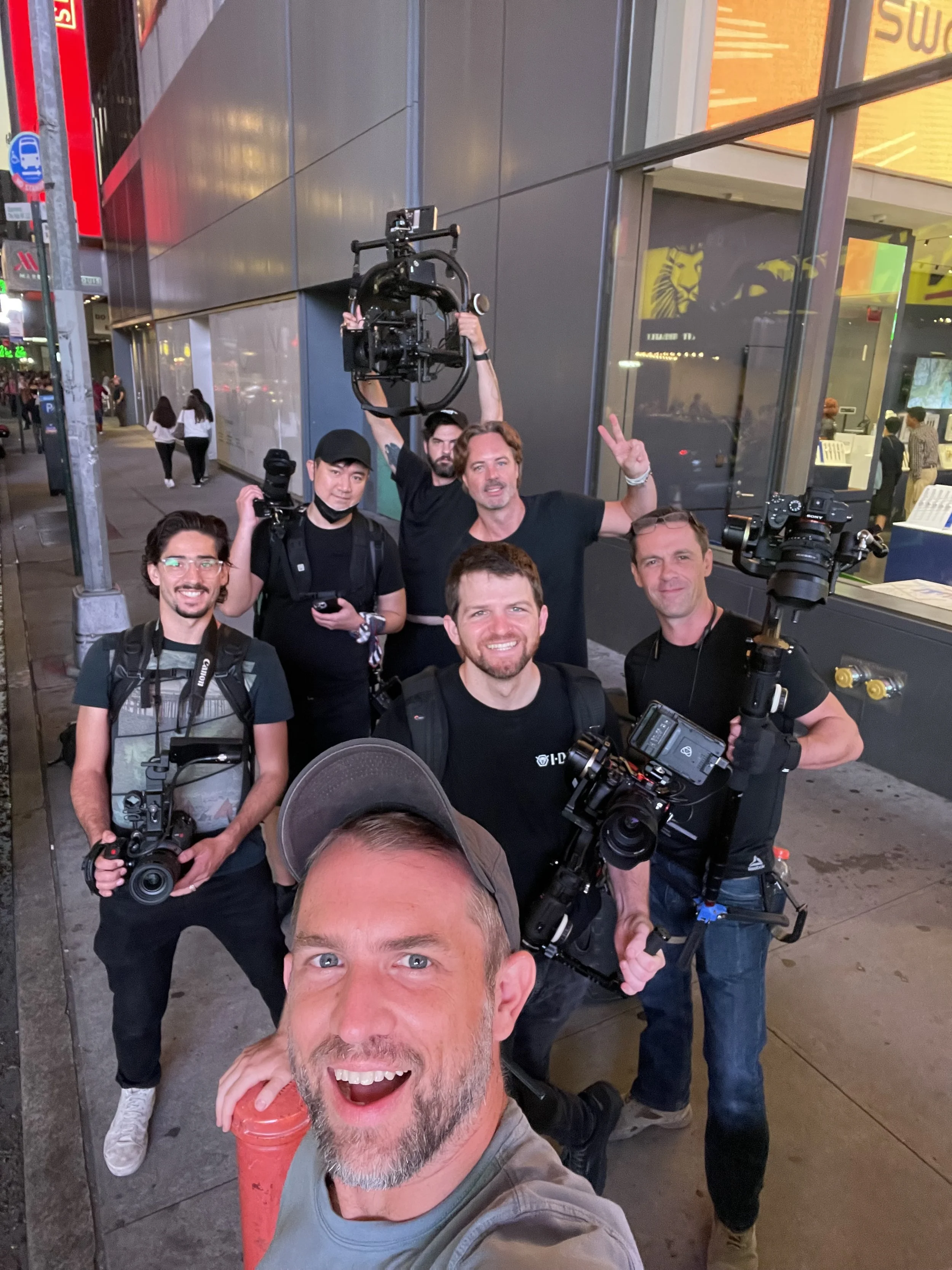 Group of seven people, some holding cameras, standing on a city sidewalk at night, with one person in the front taking a selfie.