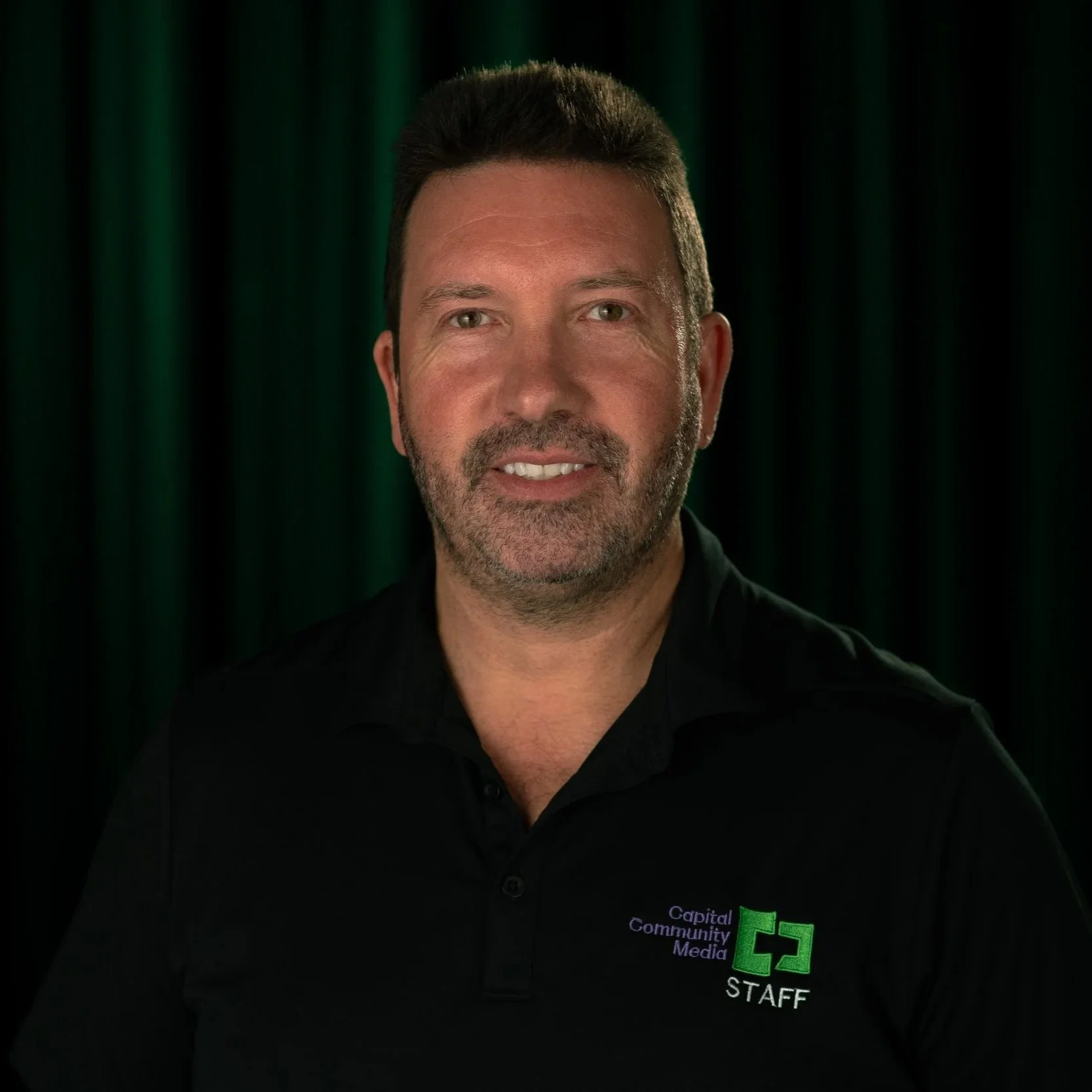 A man in black shit with Capital Community Media Staff on it with a back and Green background.