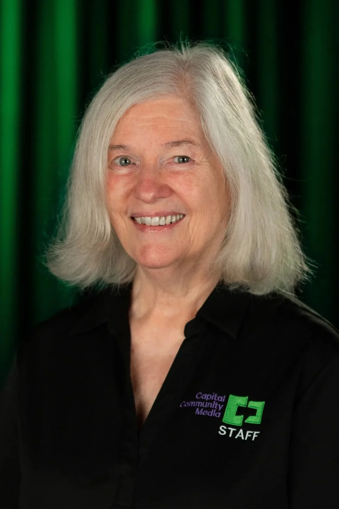 A Woman in black shit with Capital Community Media Staff on it with a back and Green background.