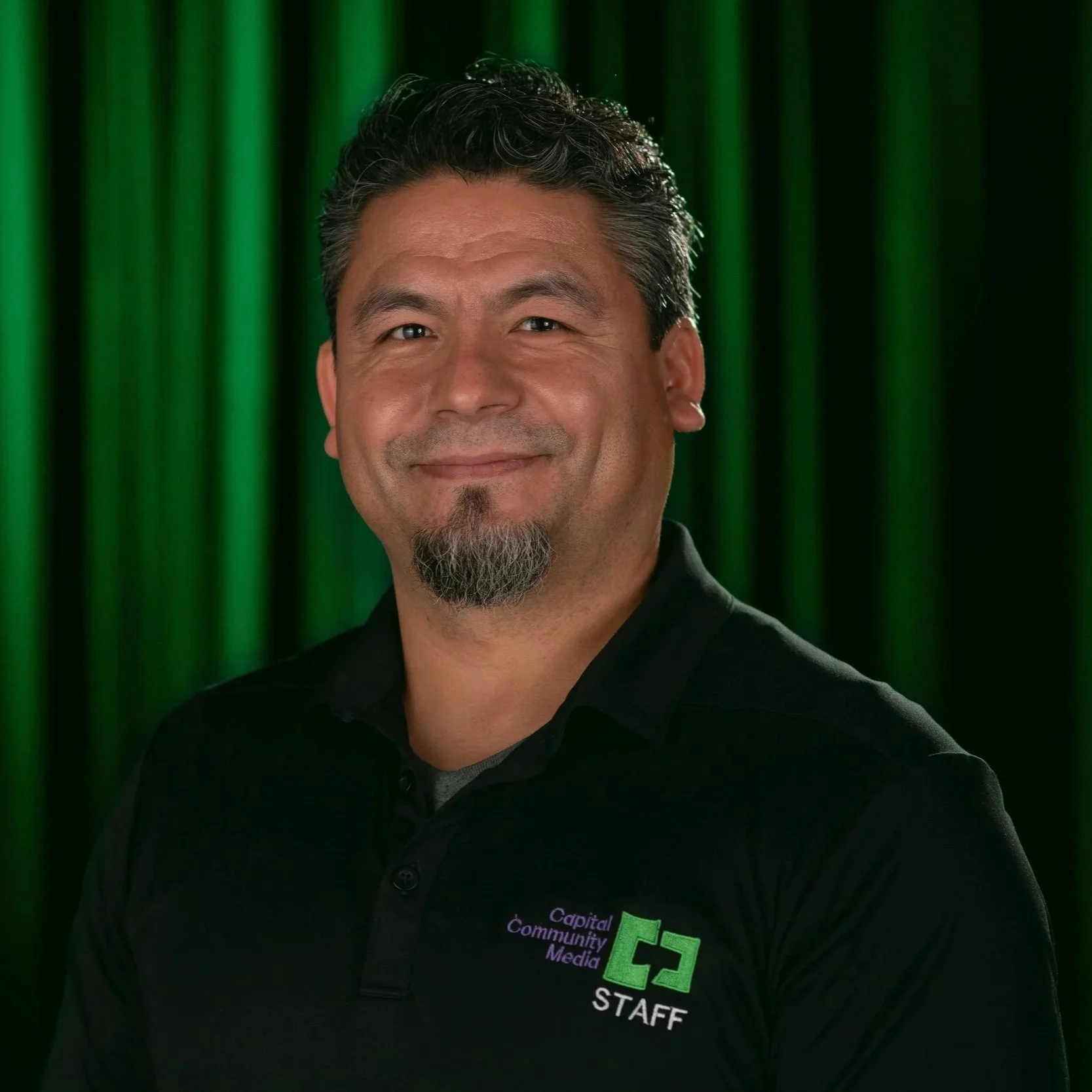 Image of a man in a Black Polo with Capital Community Media Staff with a Black background and green light.