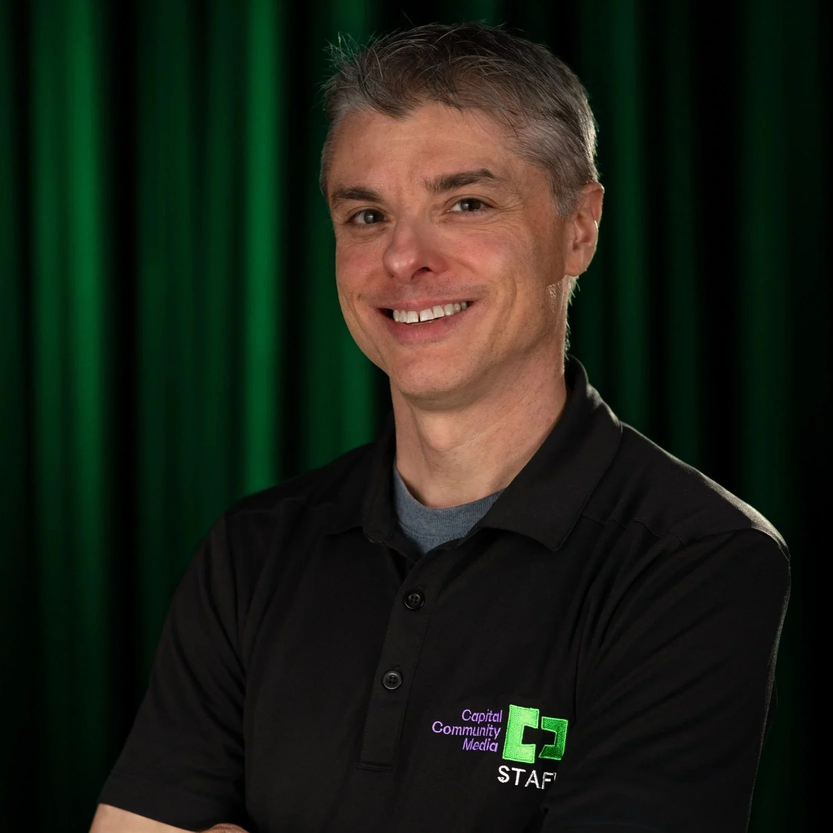 A man in black shit with Capital Community Media Staff on it with a back and Green background.