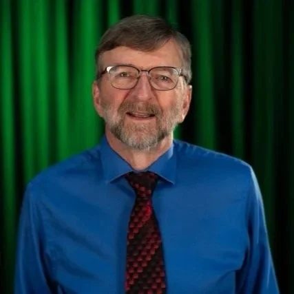 man with glasses, short brown hair, and trimmed beard, wearing a blue dress shirt and patterned dark tie, posed in front of a black with green light backdrop.
